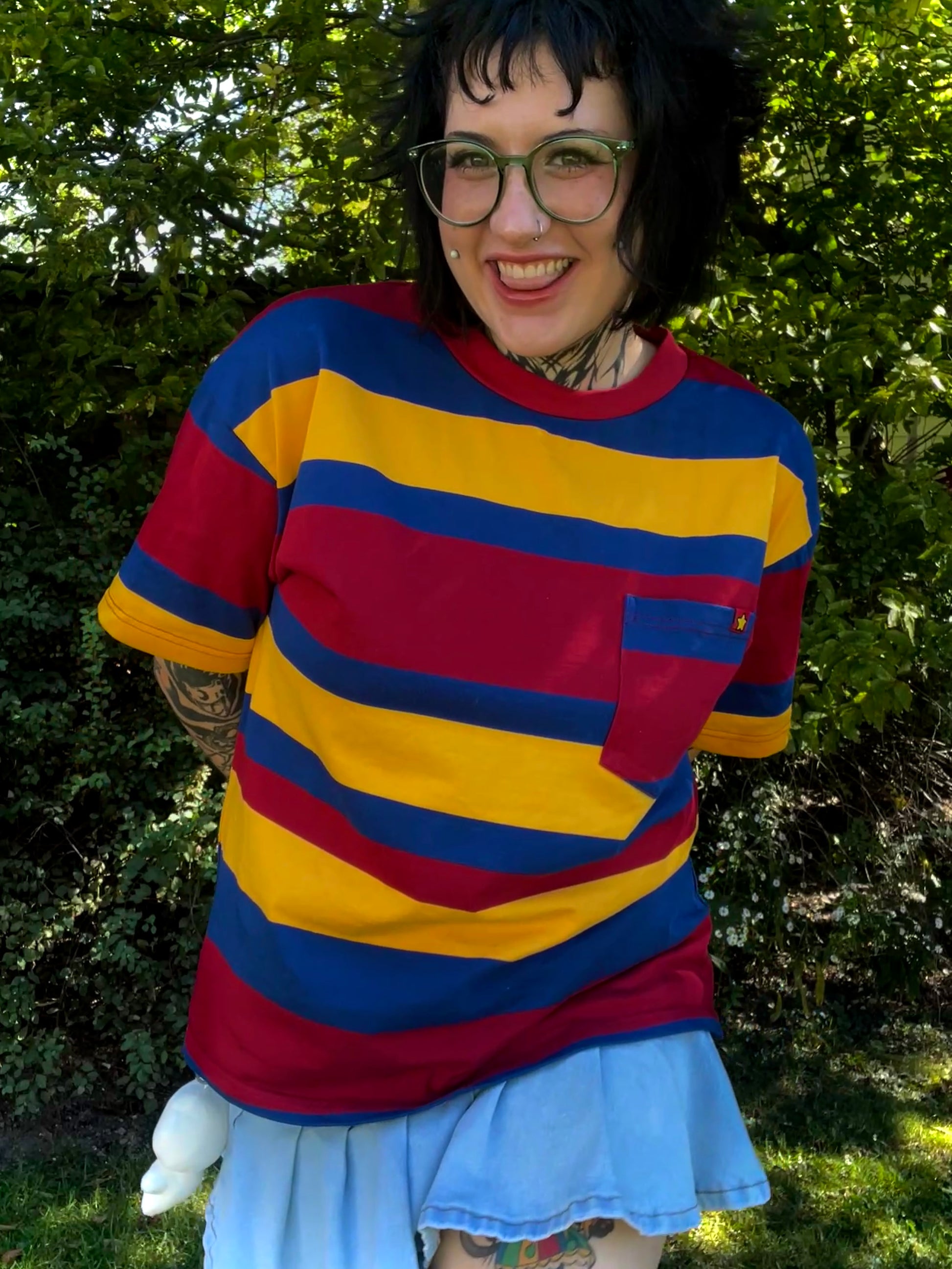 Person wearing a bright red ,yellow and blue striped shirt with a jean skirt outdoors.