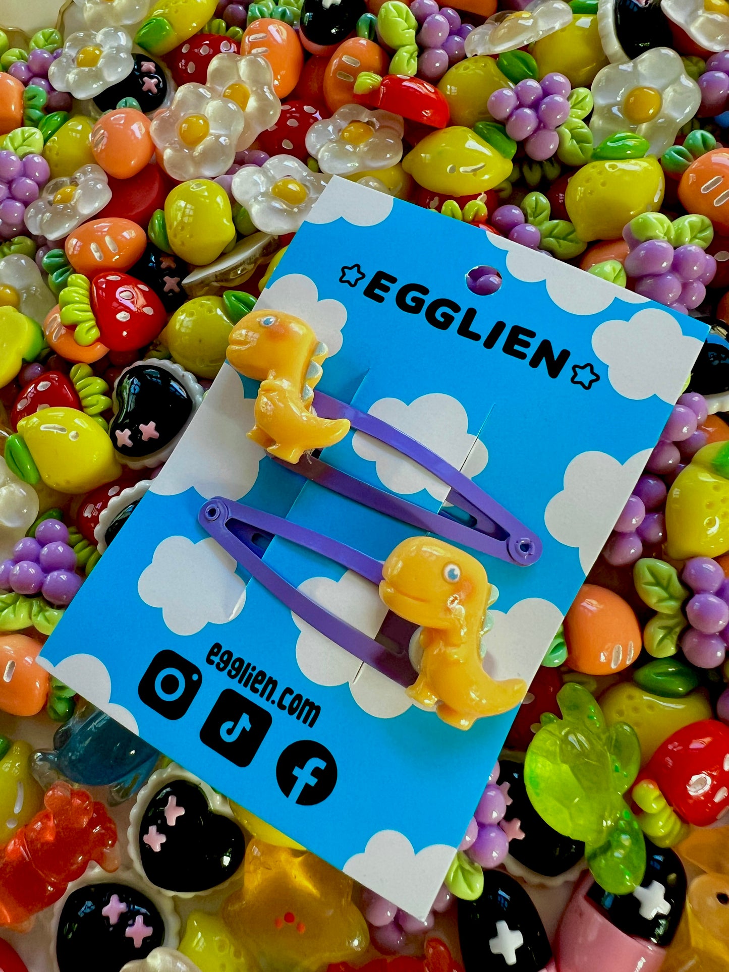 Yellow dinosaur-shaped hair clips on a blue card with 'EGGLIEN' branding, surrounded by colorful decorative resin pieces.