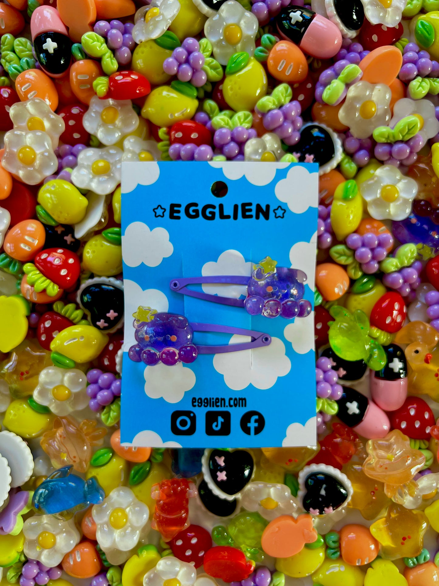 Two purple octopus with a yellow star hair clips on a blue card with 'EGGLIEN' branding, surrounded by colorful decorative resin pieces.