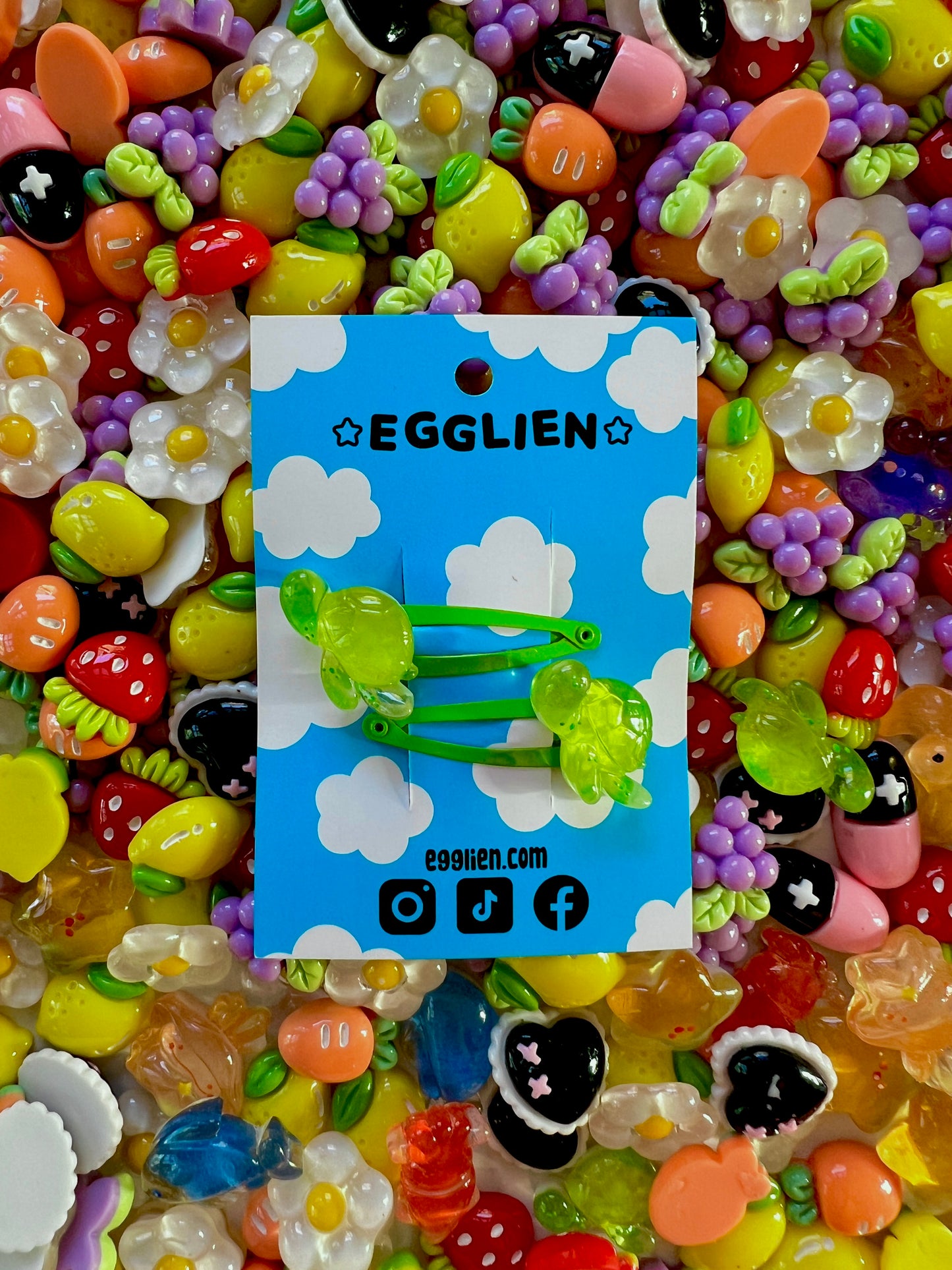 Two green turtle hair clips on a blue card with 'EGGLIEN' branding, surrounded by colorful decorative resin pieces