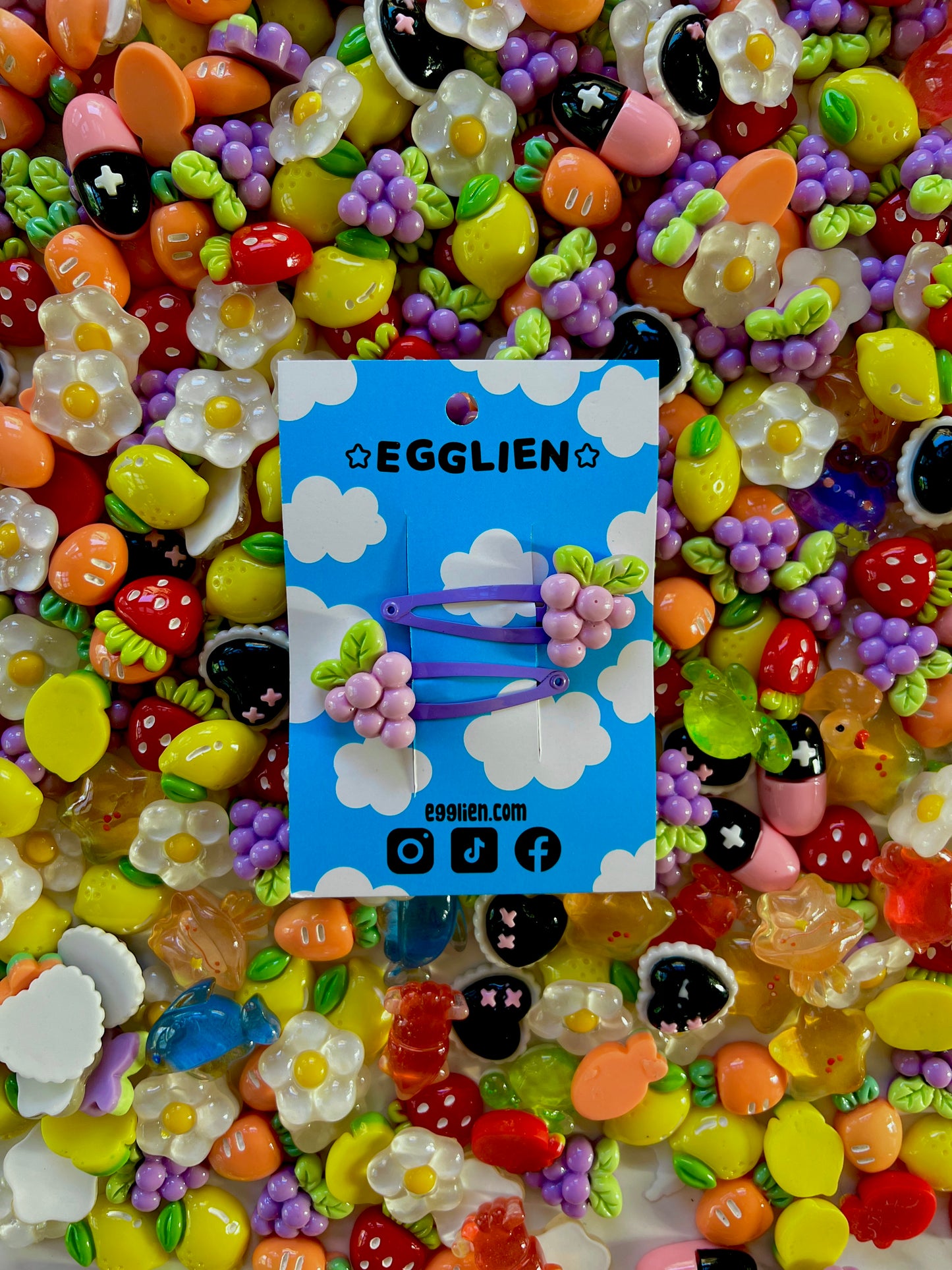Two purple grape hair clips on a blue card with 'EGGLIEN' branding, surrounded by colorful decorative resin pieces.