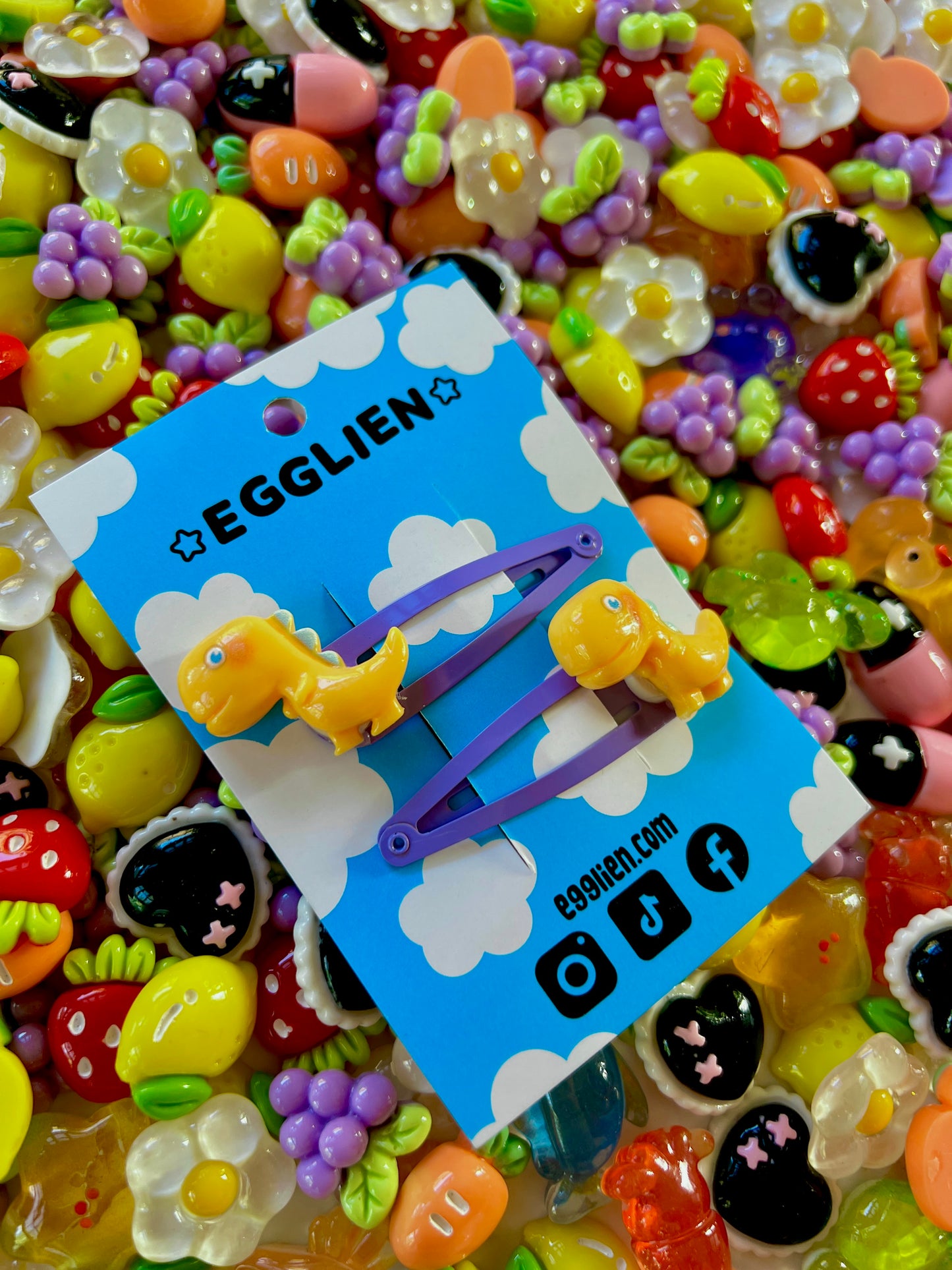 Yellow dinosaur-shaped hair clips on a blue card with 'EGGLIEN' branding, surrounded by colorful decorative resin pieces.