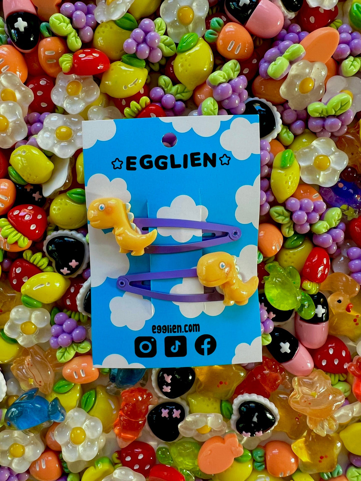 Yellow dinosaur-shaped hair clips on a blue card with 'EGGLIEN' branding, surrounded by colorful decorative resin pieces.