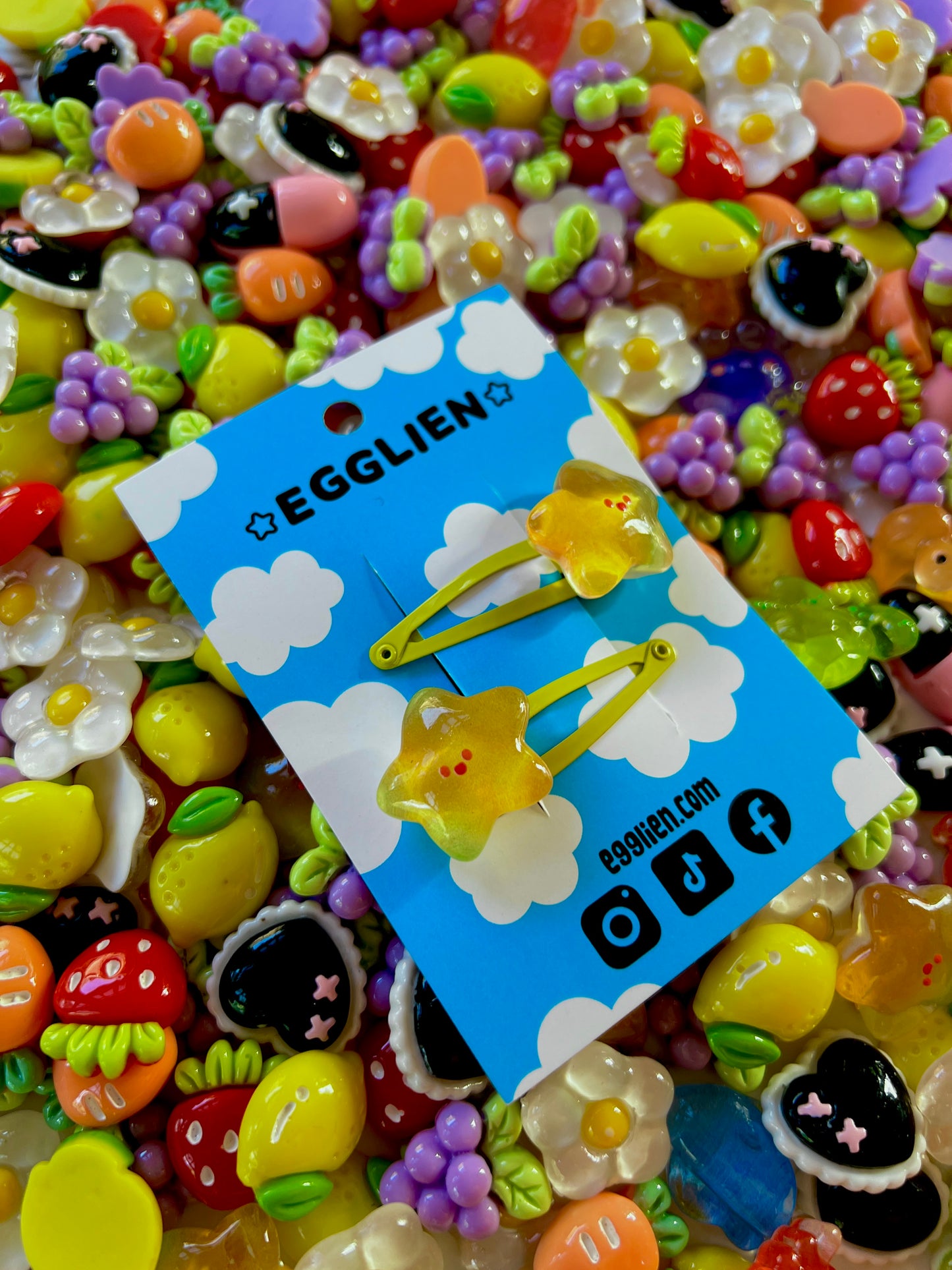 Two yellow star with happy faces hair clips on a blue card with 'EGGLIEN' branding, surrounded by colorful decorative resin pieces