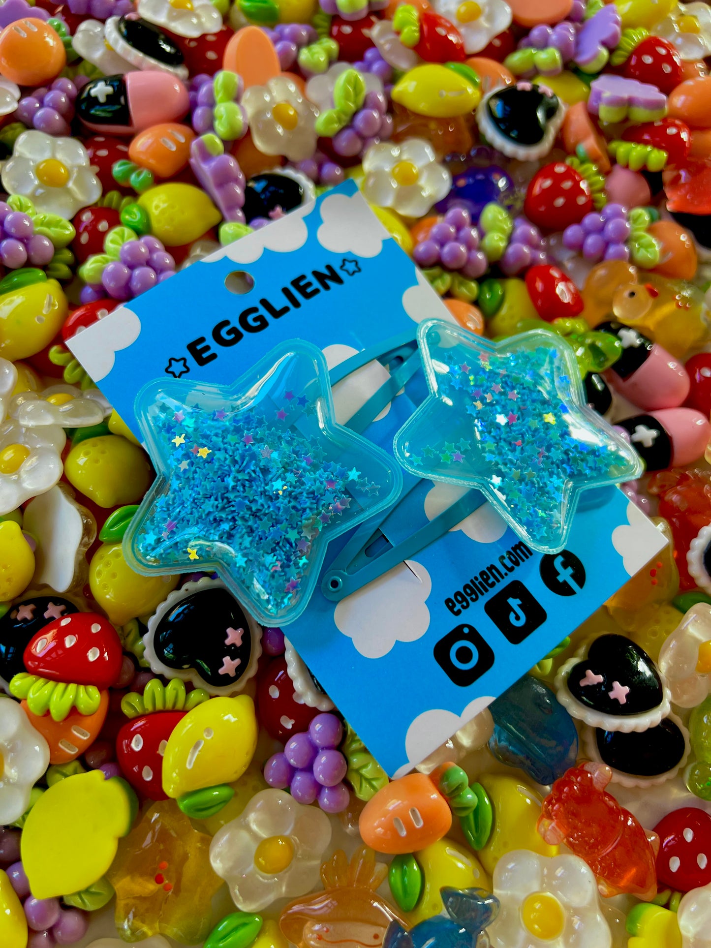 Two blue pvc stars filled with star glitter hair clips on a blue card with 'EGGLIEN' branding, surrounded by colorful decorative resin pieces