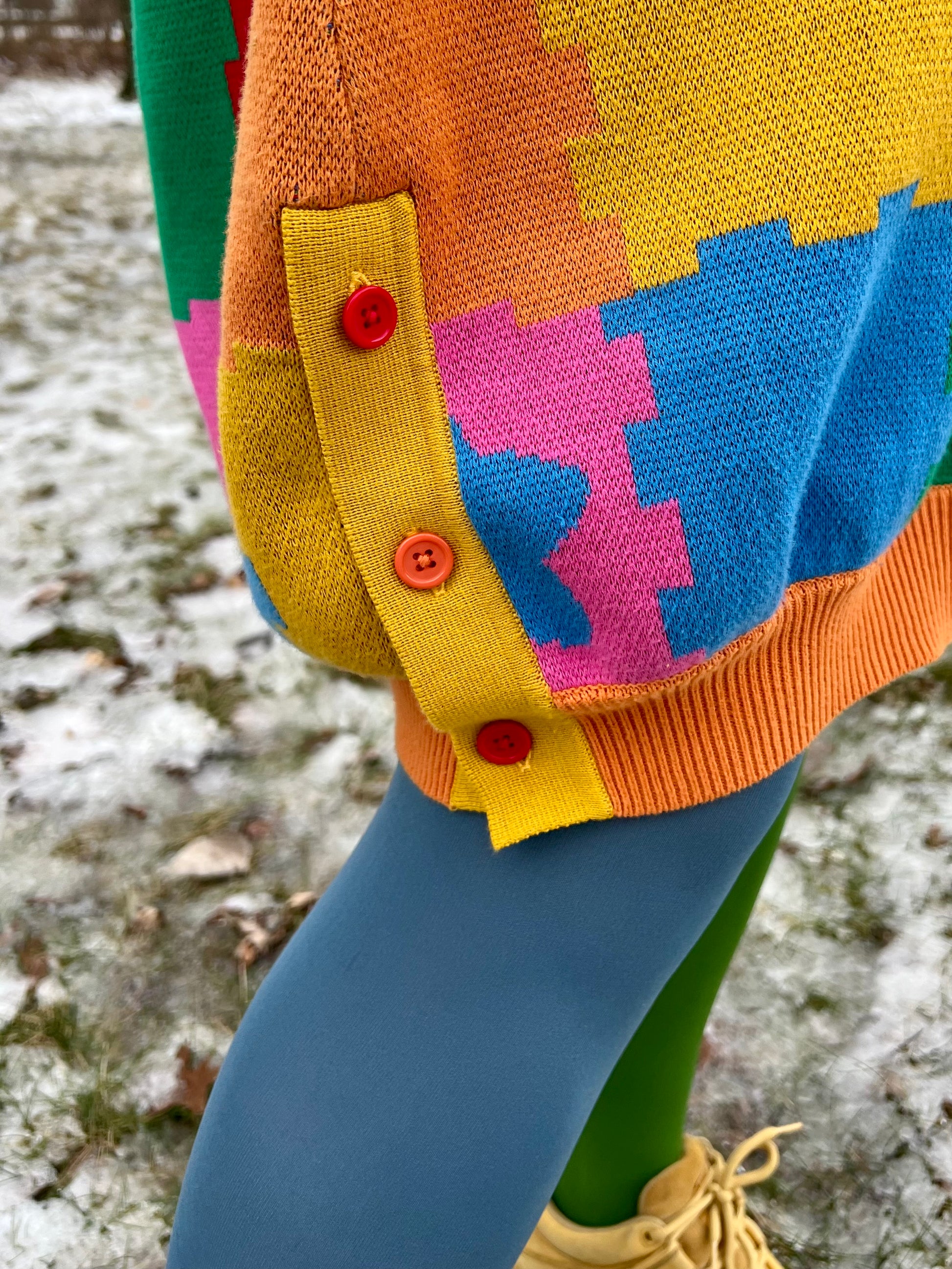 Close up image of a person wearing colorful knit design and a blue and green tights. We see the bottom orange ribbing of the dress with a yellow side cuff. The side cuff shows two red buttons and an orange button.