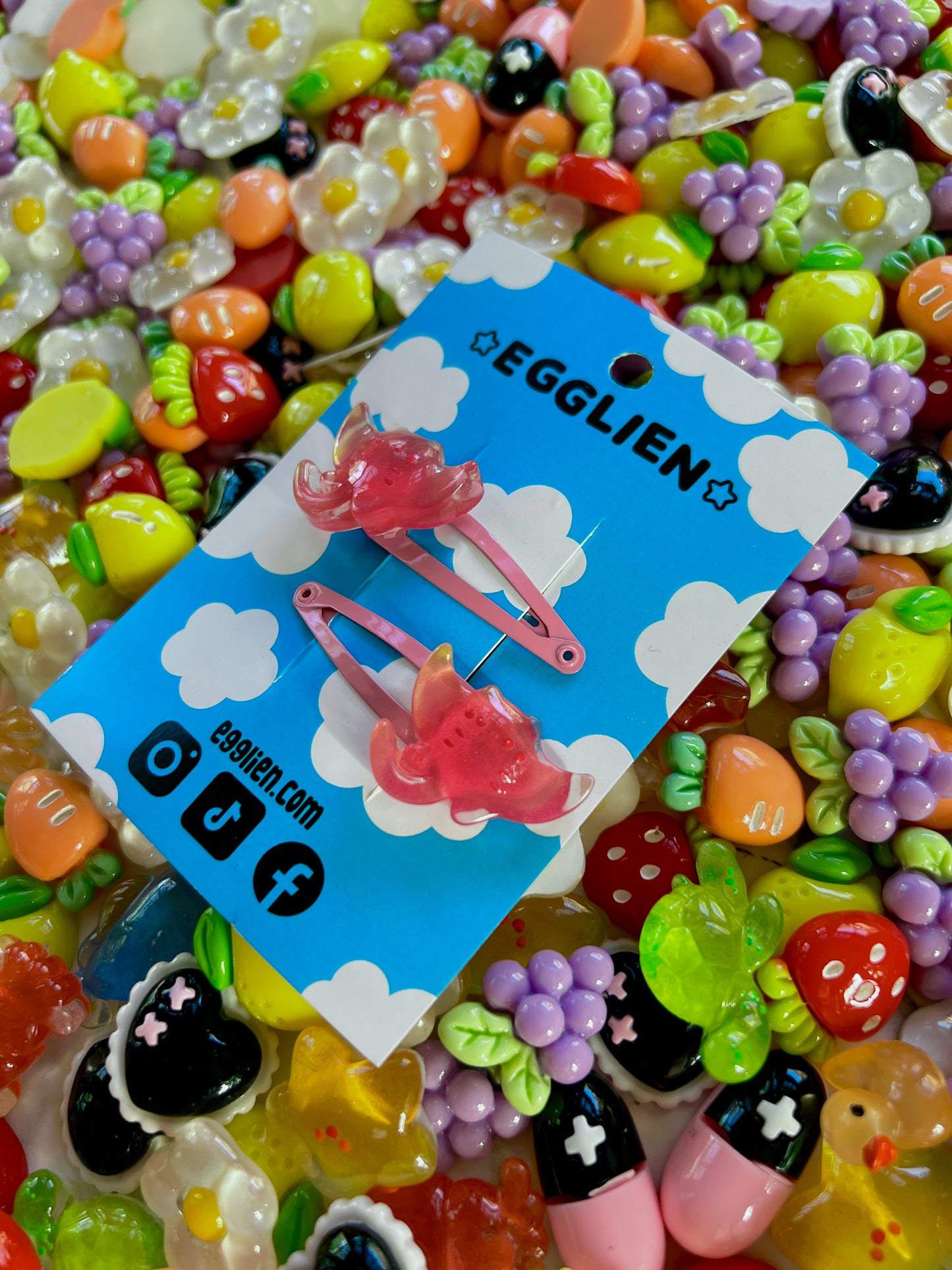 Two pink ray hair clips on a blue card with 'EGGLIEN' branding, surrounded by colorful decorative resin pieces