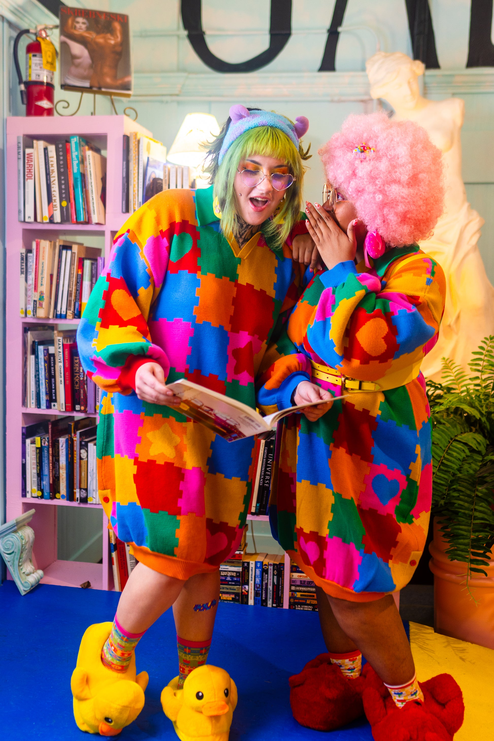 Two girls are standing in the center. One girl is wispering to the other while she reads. Both are wearing colorful sweater dresses that have the design of puzzle play mats on them. Behind them is a bookshelf filled with books, a plant, and a large sculpture of a woman.