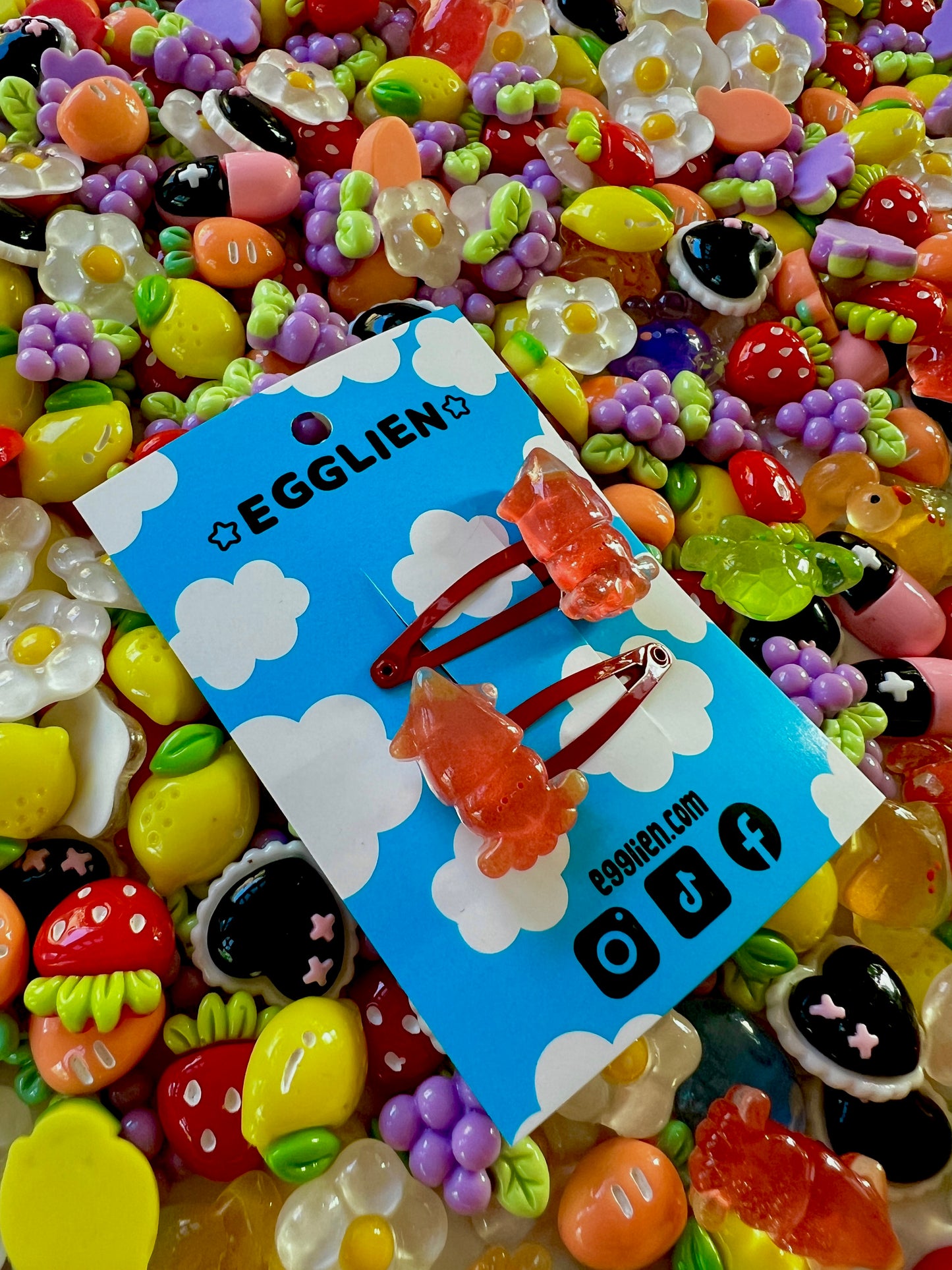 Two red squid hair clips on a blue card with 'EGGLIEN' branding, surrounded by colorful decorative resin pieces