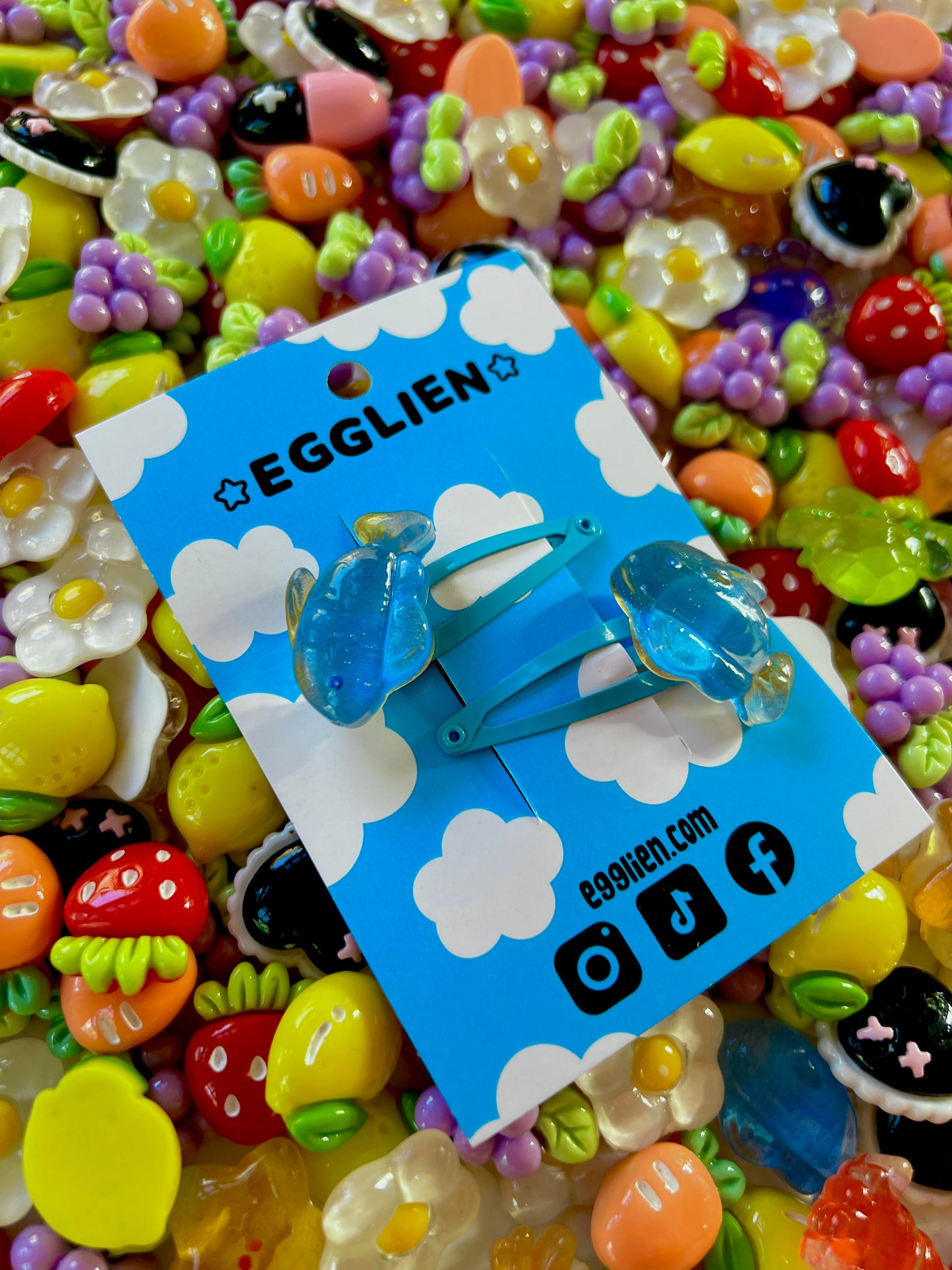 Two blue fish hair clips on a blue card with 'EGGLIEN' branding, surrounded by colorful decorative resin pieces