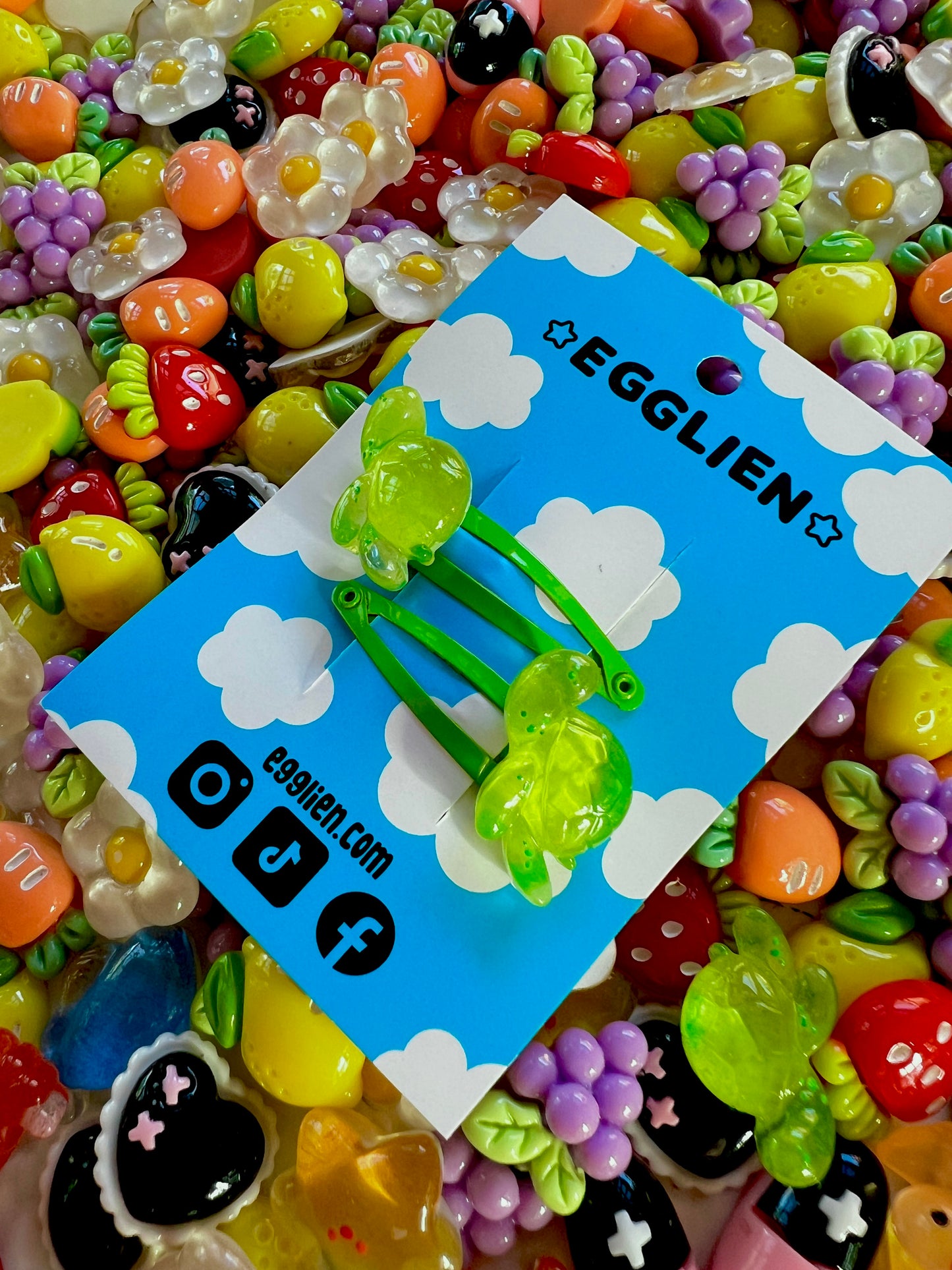 Two green turtle hair clips on a blue card with 'EGGLIEN' branding, surrounded by colorful decorative resin pieces