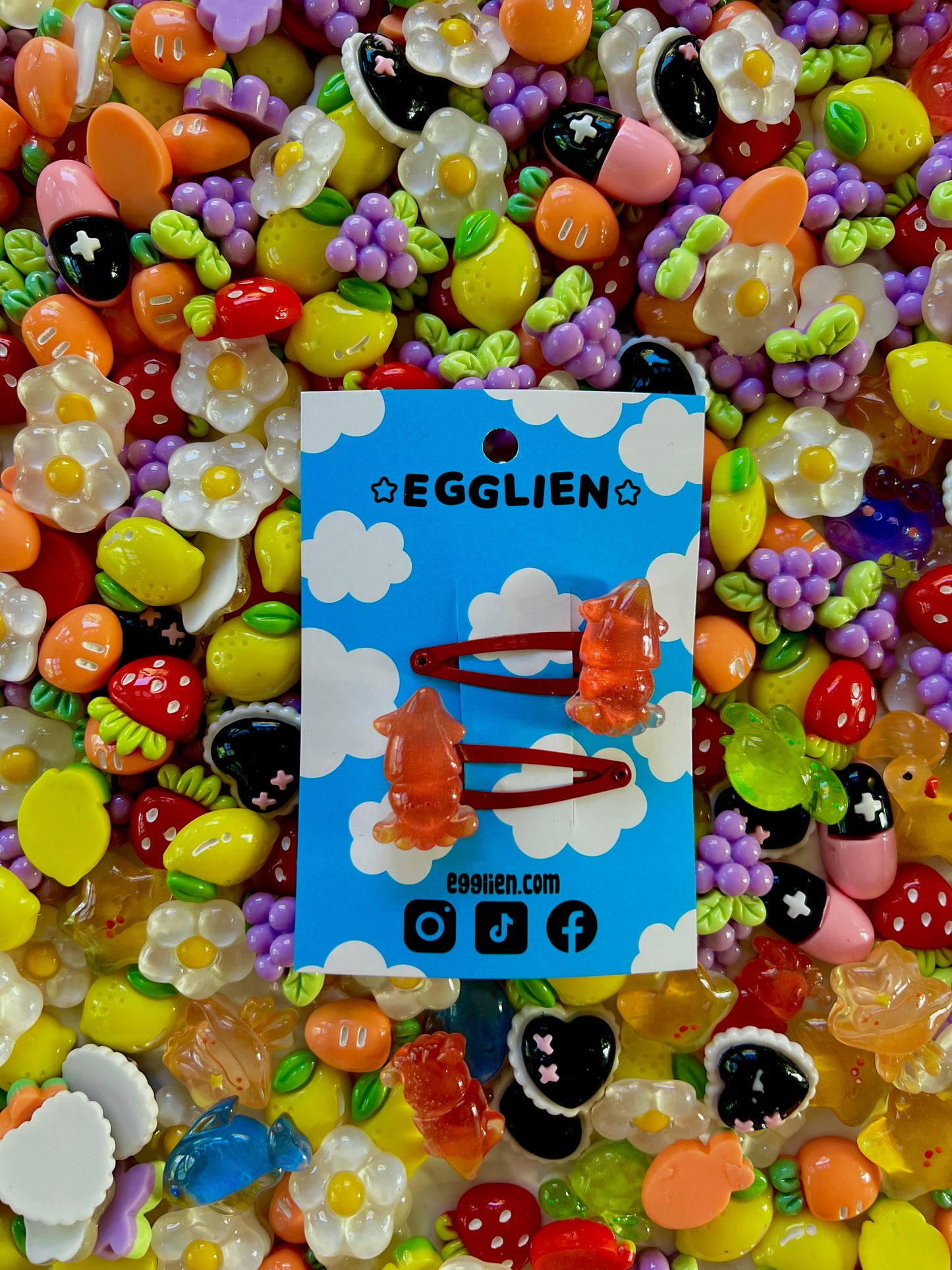 Two red squid hair clips on a blue card with 'EGGLIEN' branding, surrounded by colorful decorative resin pieces