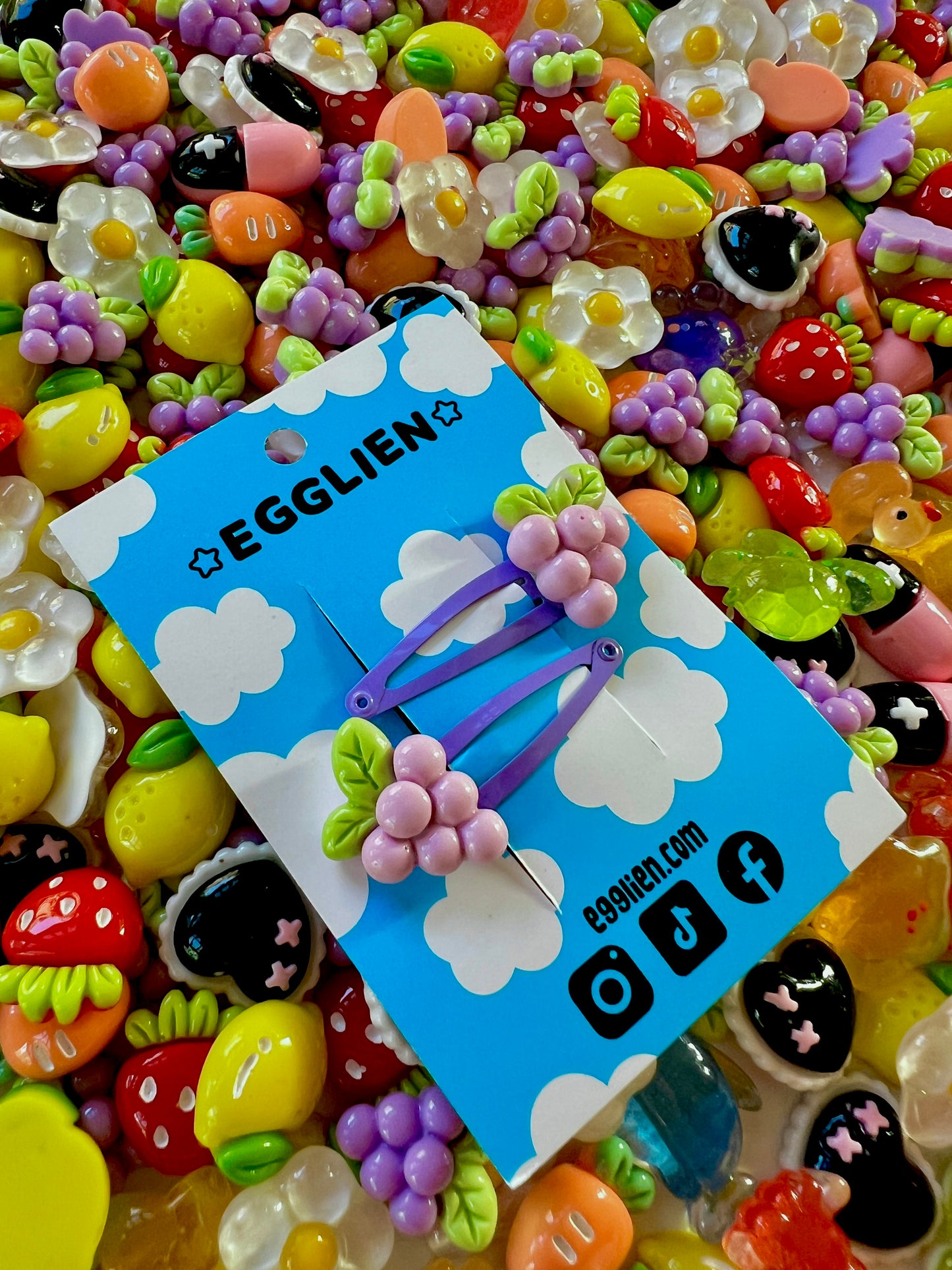 Two purple grape hair clips on a blue card with 'EGGLIEN' branding, surrounded by colorful decorative resin pieces.