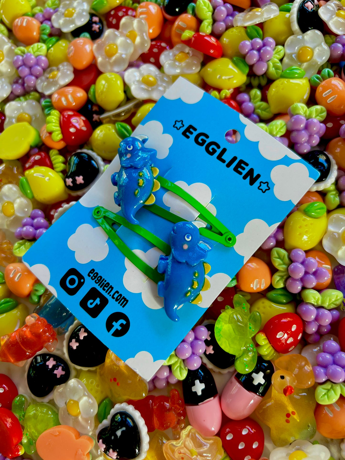 Dark Blue dinosaur-shaped hair clips on a blue card with 'EGGLIEN' branding, surrounded by colorful decorative resin pieces.