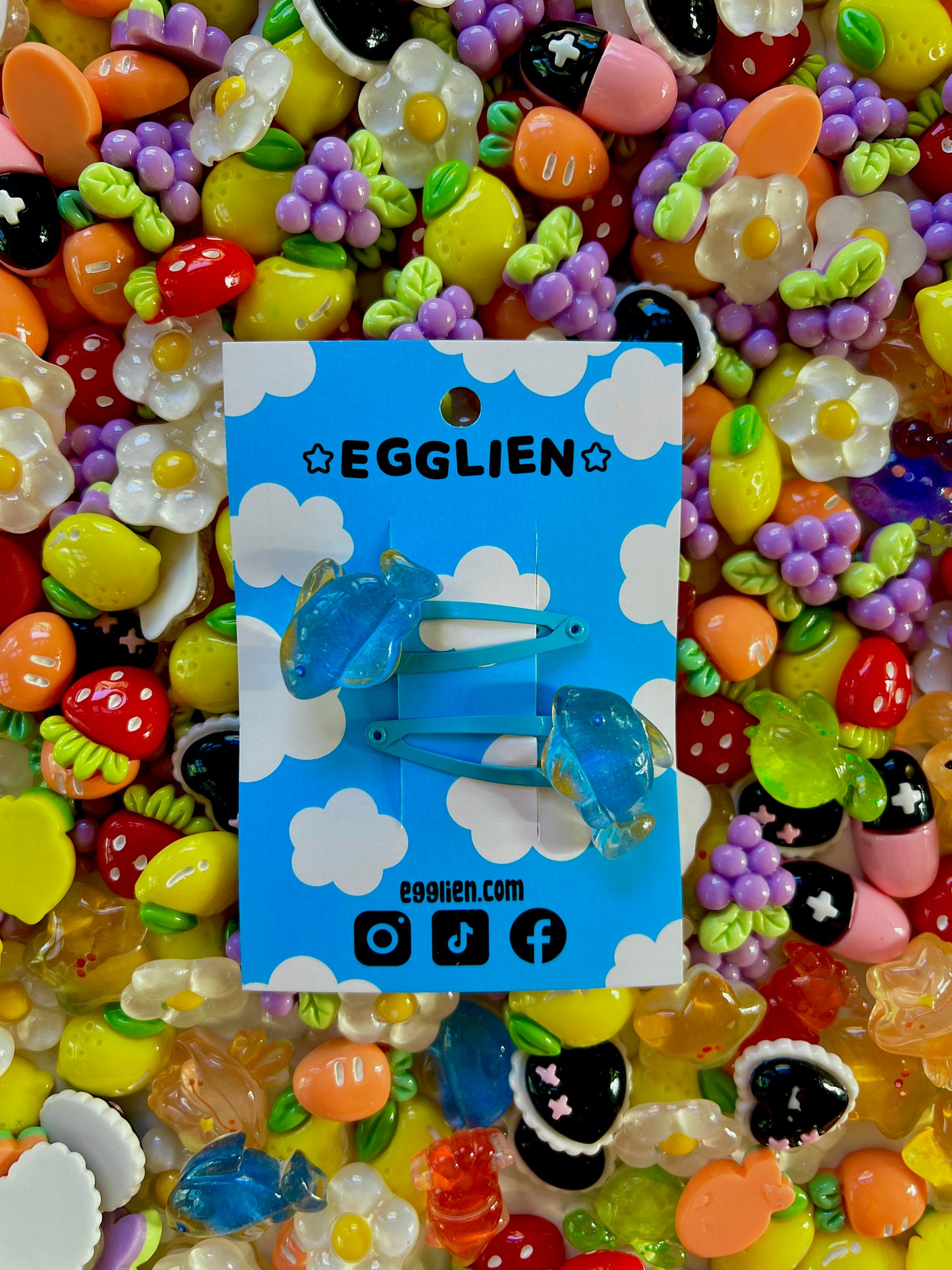 Two blue fish hair clips on a blue card with 'EGGLIEN' branding, surrounded by colorful decorative resin pieces