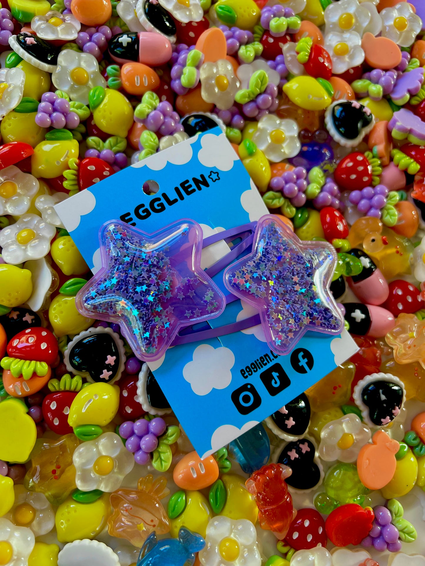 Two purple pvc stars filled with star glitter hair clips on a blue card with 'EGGLIEN' branding, surrounded by colorful decorative resin pieces
