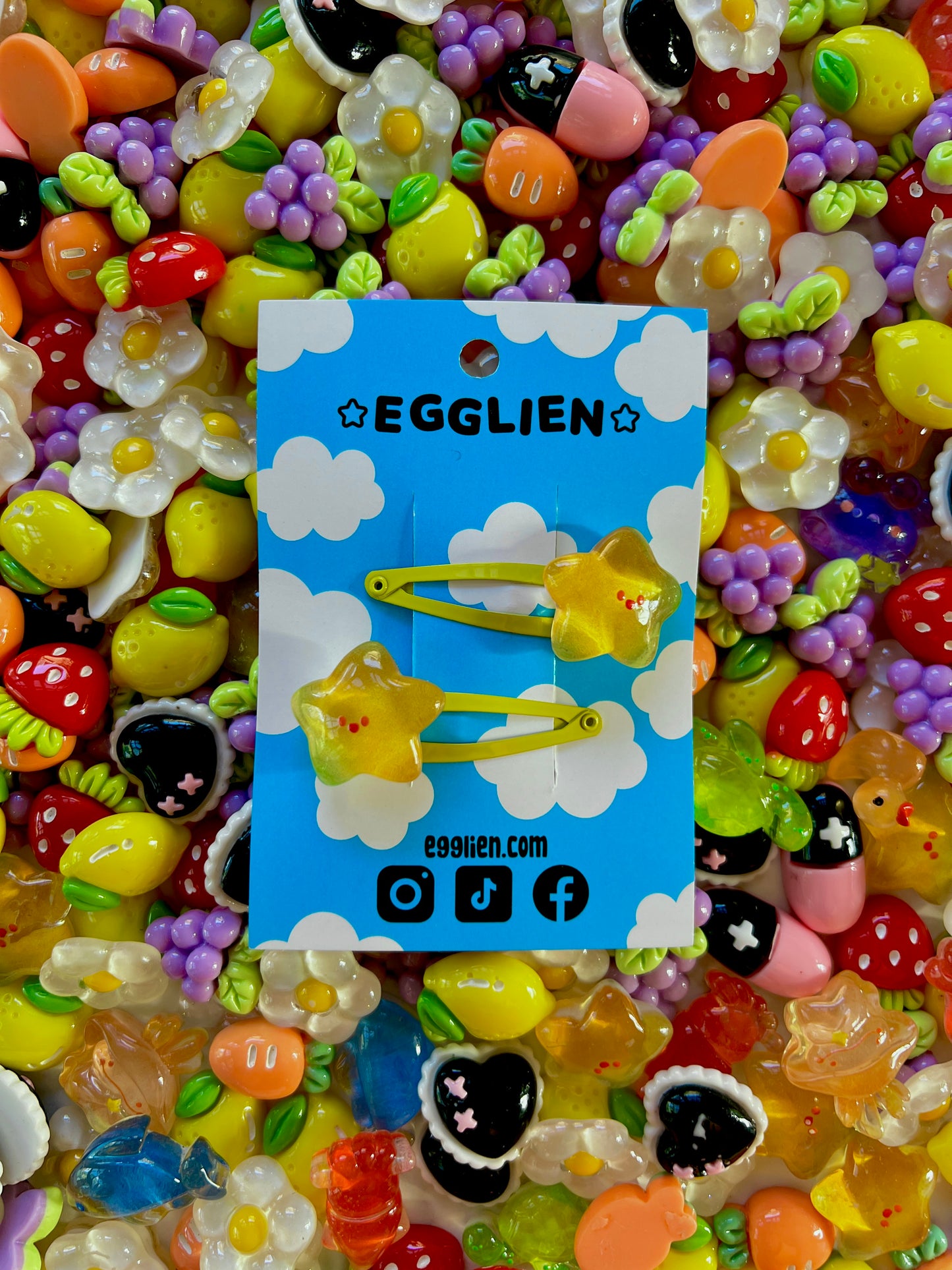 Two yellow star with happy faces hair clips on a blue card with 'EGGLIEN' branding, surrounded by colorful decorative resin pieces