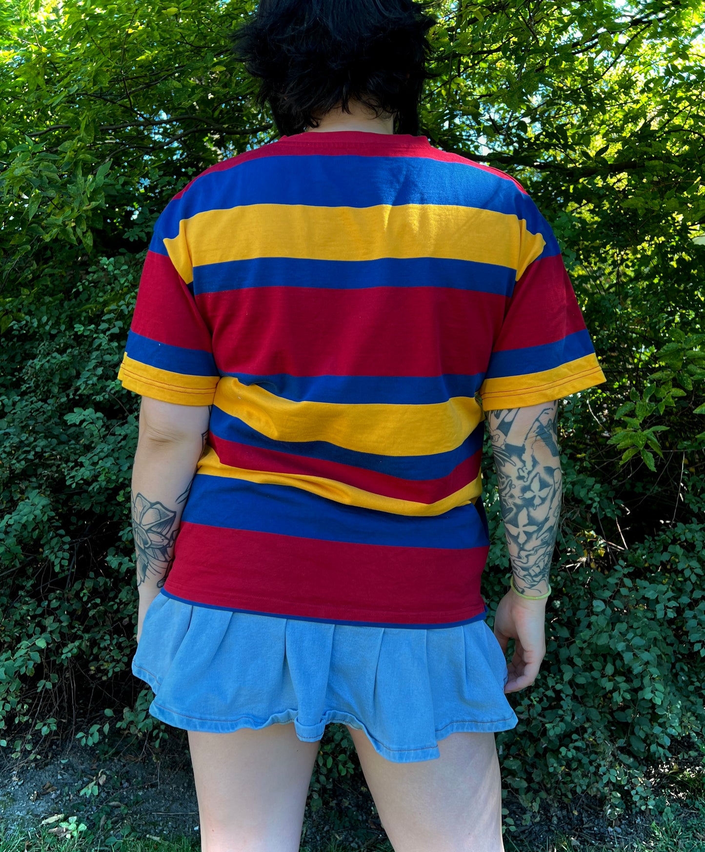 Person wearing a red, yellow, and blue striped shirt with a blue skirt outdoors with back facing the camera showing the back of the shirt