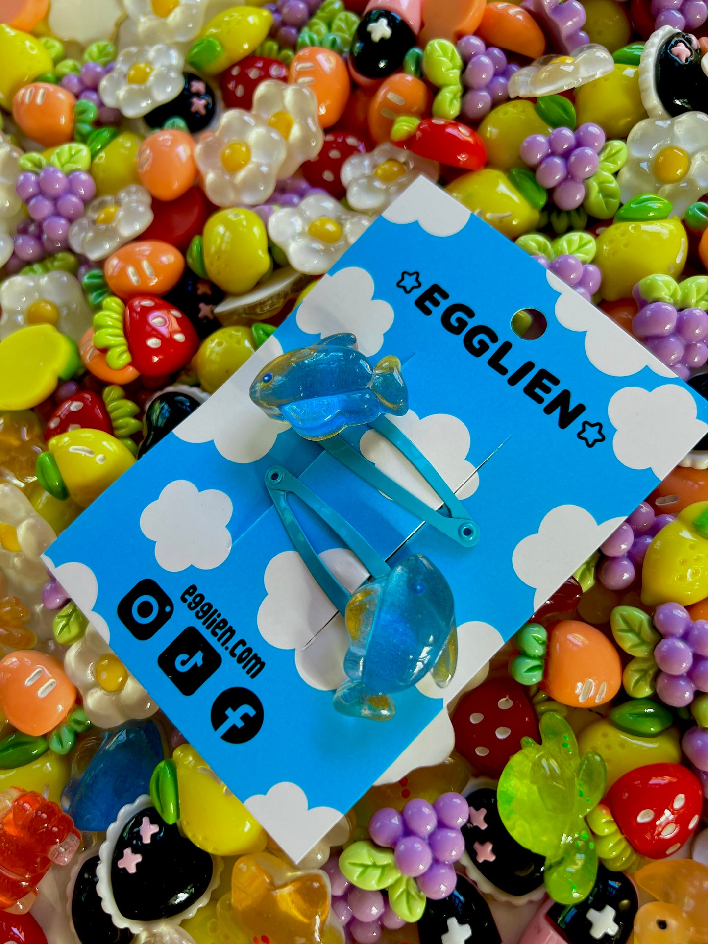 Two blue fish hair clips on a blue card with 'EGGLIEN' branding, surrounded by colorful decorative resin pieces