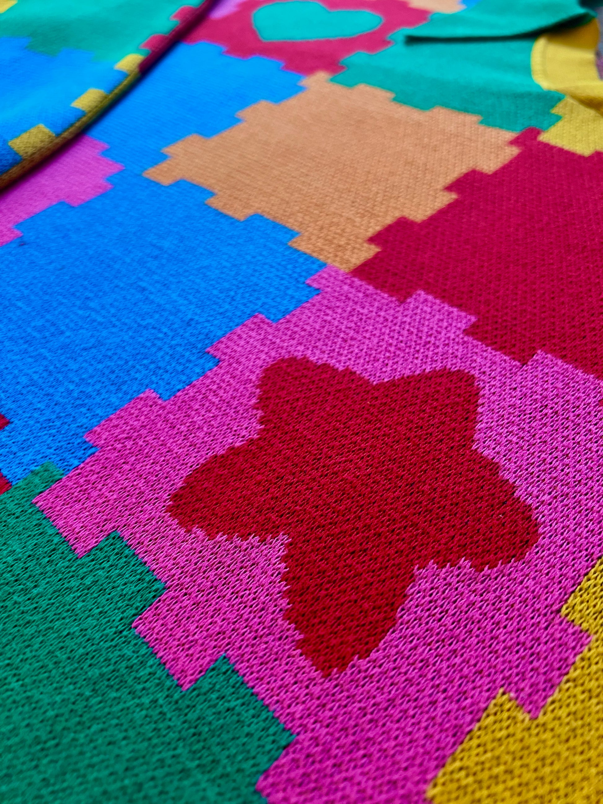 Colorful knit sweater dress is laying on the ground.The dress is bright blue, green, orange, pink, yellow and red with the design of puzzle playmats.  Close up we see the texture of the knitted fibers.