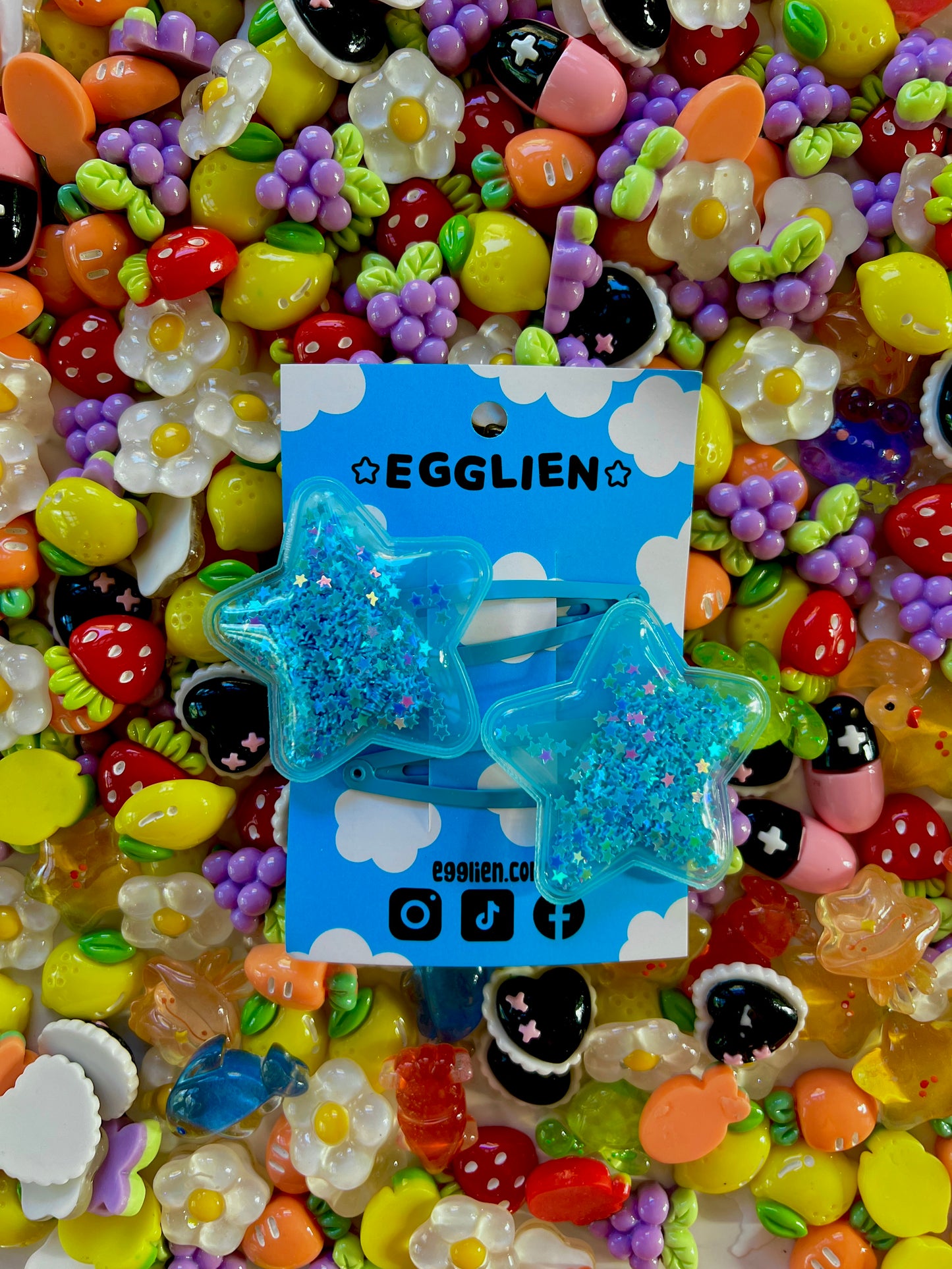 Two blue pvc stars filled with star glitter hair clips on a blue card with 'EGGLIEN' branding, surrounded by colorful decorative resin pieces