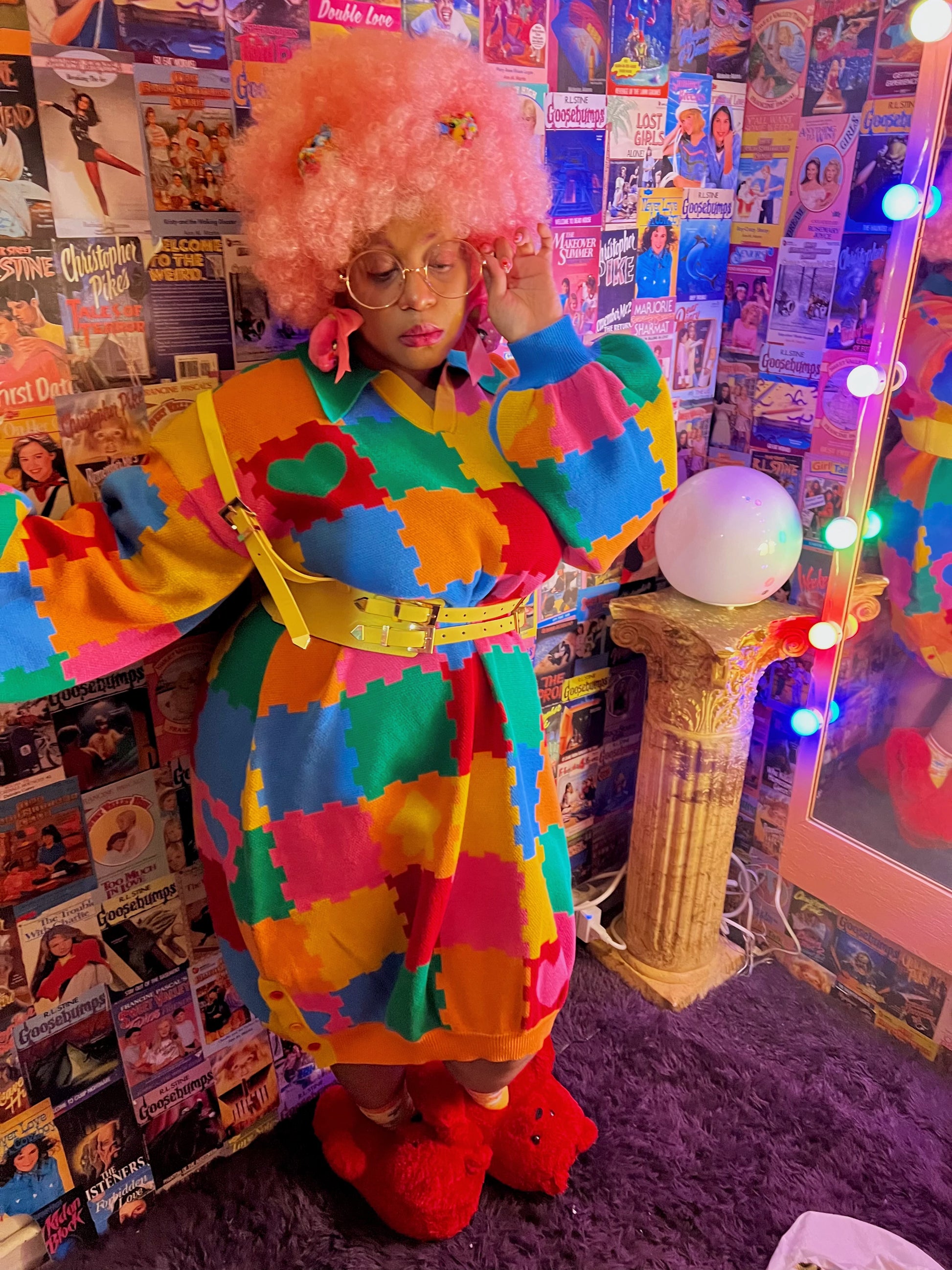 Girl is standing facing away from a mirror and looking down at the ground. She is wearing a knit sweater dress with a colorful puzzle playmat design. she is also wearing a yellow belt, pink hair, and red bear slippers. Behind her is a collage of miscellaneous retro book covers.