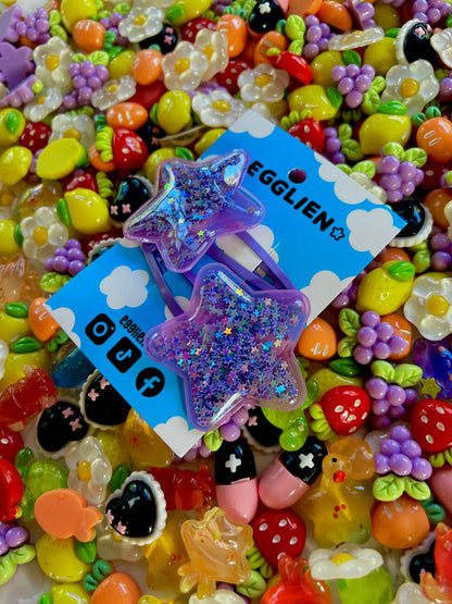 Two purple pvc stars filled with star glitter hair clips on a blue card with 'EGGLIEN' branding, surrounded by colorful decorative resin pieces