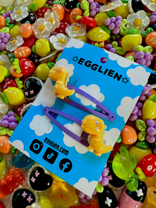 Yellow dinosaur-shaped hair clips on a blue card with 'EGGLIEN' branding, surrounded by colorful decorative resin pieces.