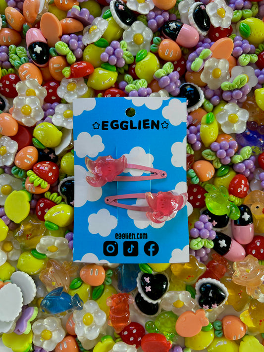 Two pink ray hair clips on a blue card with 'EGGLIEN' branding, surrounded by colorful decorative resin pieces