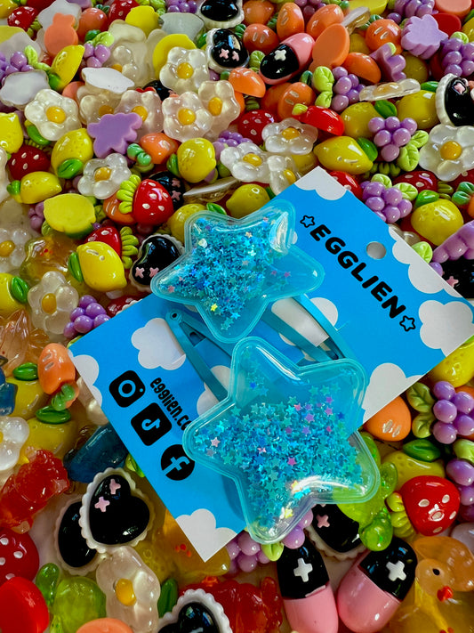 Two blue pvc stars filled with star glitter hair clips on a blue card with 'EGGLIEN' branding, surrounded by colorful decorative resin pieces