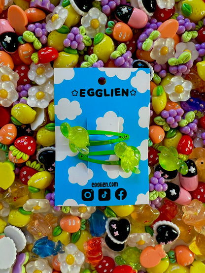 Two green turtle hair clips on a blue card with 'EGGLIEN' branding, surrounded by colorful decorative resin pieces