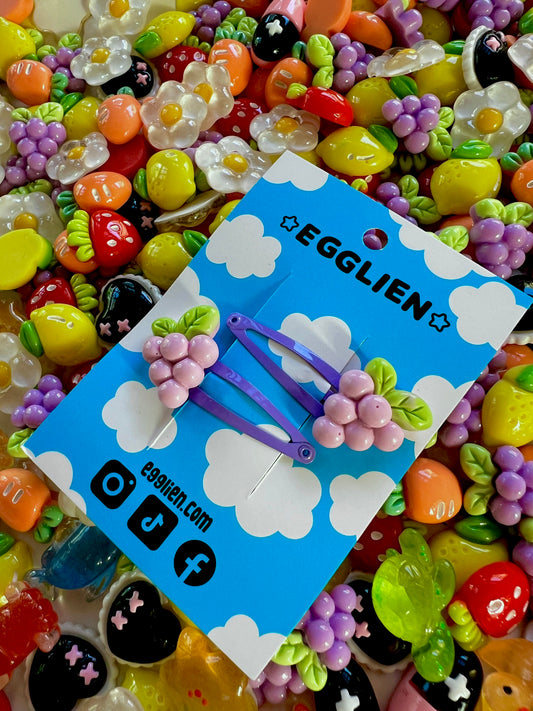 Two purple grape hair clips on a blue card with 'EGGLIEN' branding, surrounded by colorful decorative resin pieces.