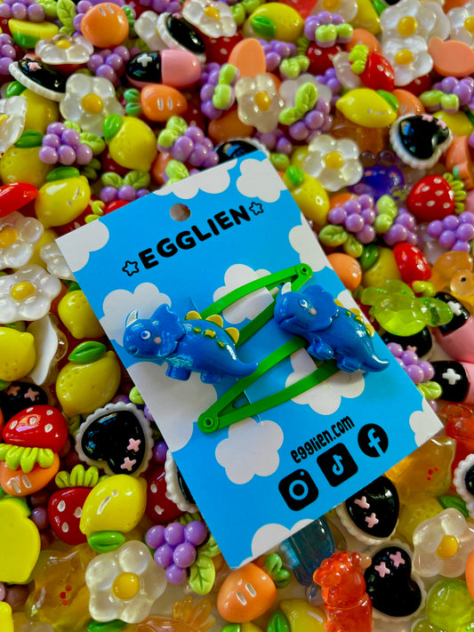 Dark Blue dinosaur-shaped hair clips on a blue card with 'EGGLIEN' branding, surrounded by colorful decorative resin pieces.