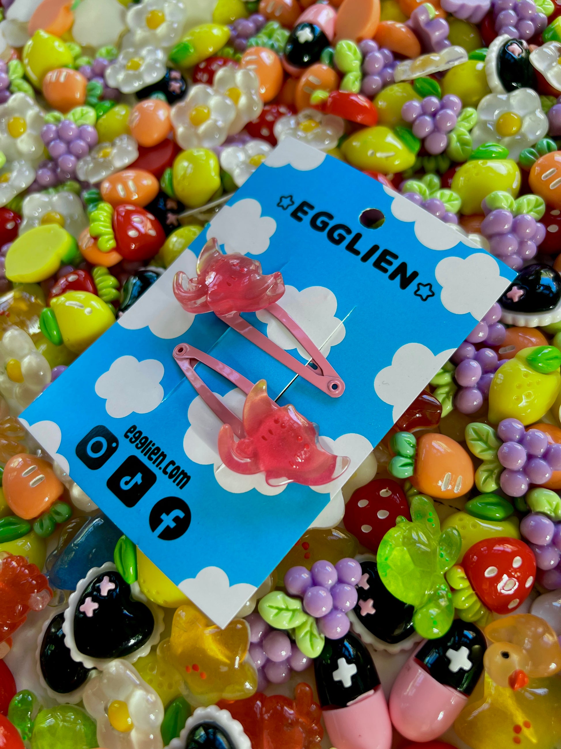 Two pink ray hair clips on a blue card with 'EGGLIEN' branding, surrounded by colorful decorative resin pieces