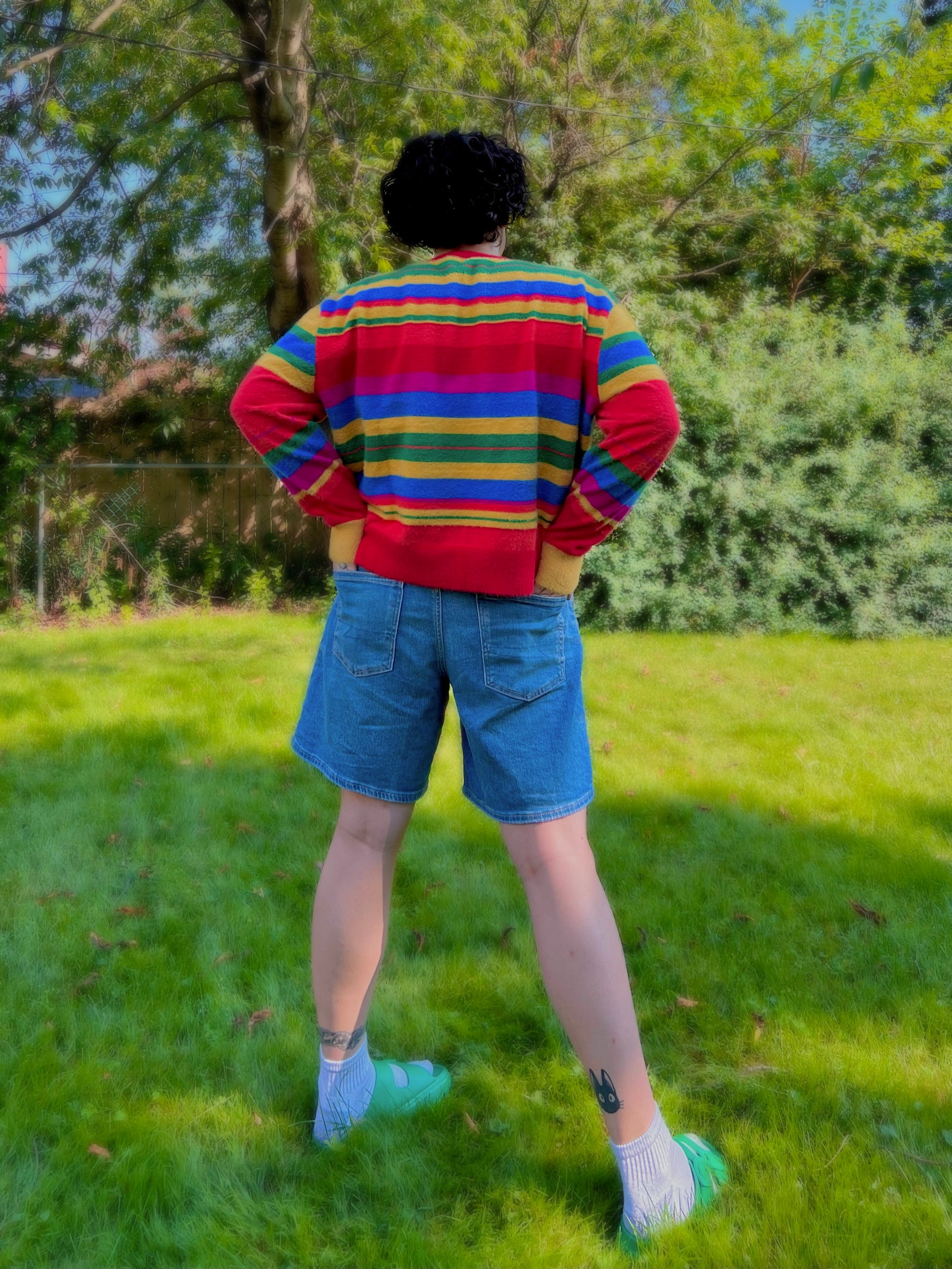 Person wearing a colorful striped sweater and denim shorts standing in a grassy area with trees.