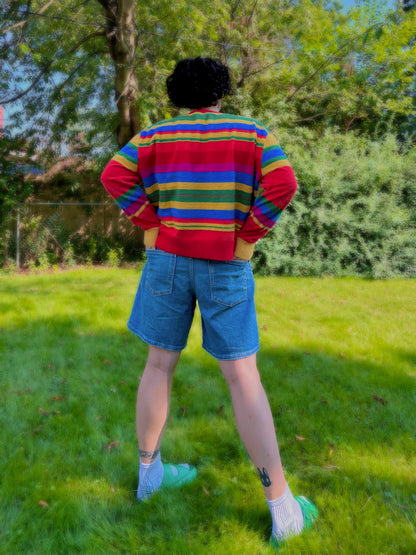 Person wearing a colorful striped sweater and denim shorts standing in a grassy area with trees.