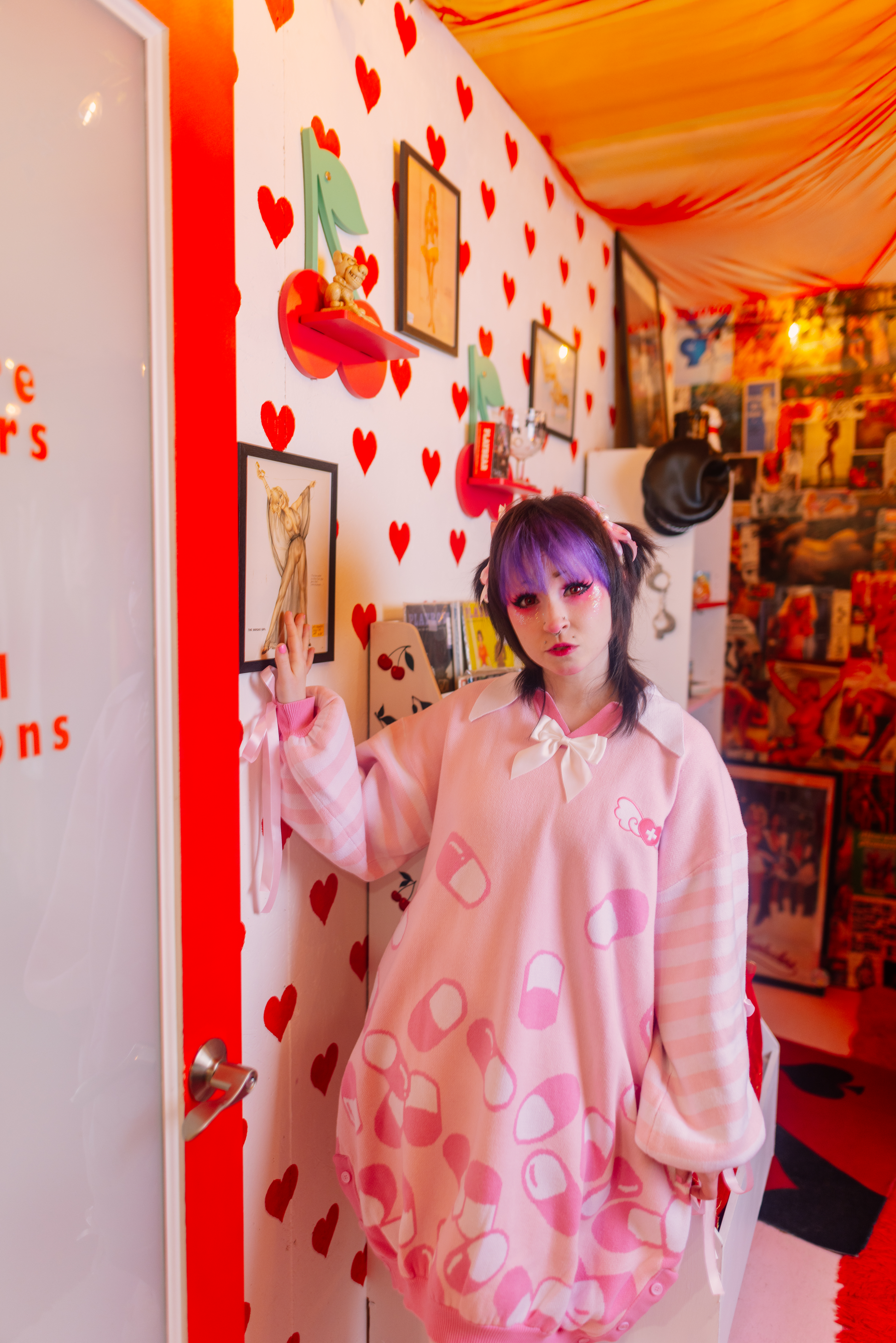 Girl is standing in room with one hand resting against the wall. She is wearing a pastel pink sweater dress with darker pink and white capsules on it with a pastel pink and white striped sleeves. Pastel pink ribbons are peaking out from the sleeves. In the background is room with cherry shelves, red heart wallpaper and pictures of women on the wall.