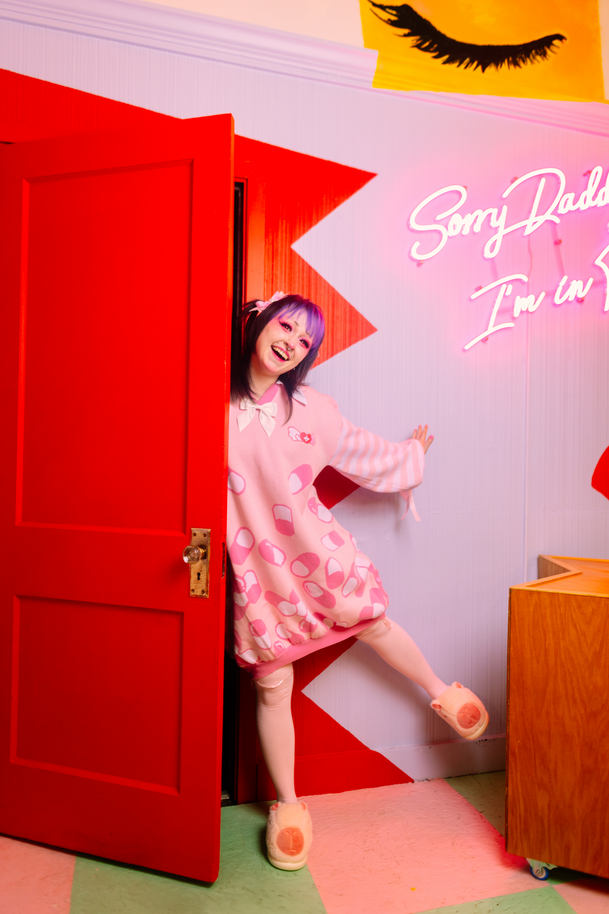 Girl is playfully opening a red door with three quarters of her body peaking out. She is wearing a pastel pink sweater dress with darker pink and white capsules on it with a pastel pink and white striped sleeves. In the background is picture of a closed eye showing eyelashes and an LED sign that reads "Sorry Daddy.. I'm In.." half of the sign is cut off.