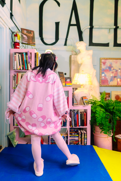 Girl standing with her back facing the camera and pulling on the sides of her sweater dress to show the back design on it. She is wearing a pastel pink sweater dress with darker pink and white capsules on it with a pastel pink and white striped sleeves. There is a heart with a white cross in it with wings coming out of the side on the shoulders part of the sweater dress. In the background is a bookshelf filled with books and big porcelain sculpture of a woman.
