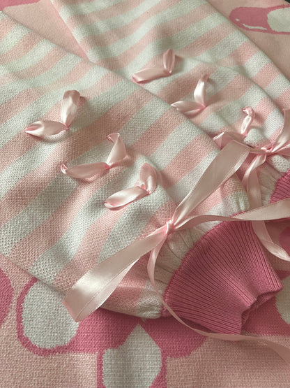 The pastel pink and white sleeves of a knit cardigan are shown close up. Showcasing the darker pink cuffs along with the pastel pink ribbons that are installed in each of the sleeves. The ribbon is shaped in three hearts that get closer to the cuffs where they combine to create a big pastel pink bow.