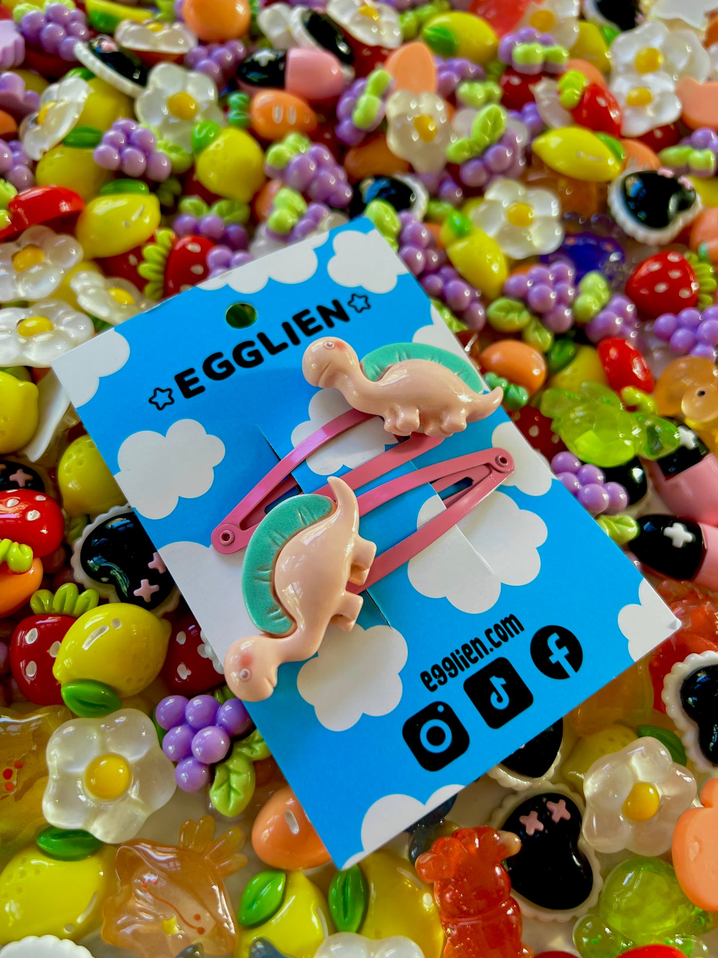 Two peach dinosaurs on bright pink hair-clips on an 'EGGLIEN' card in the center surrounded by colorful decorative items