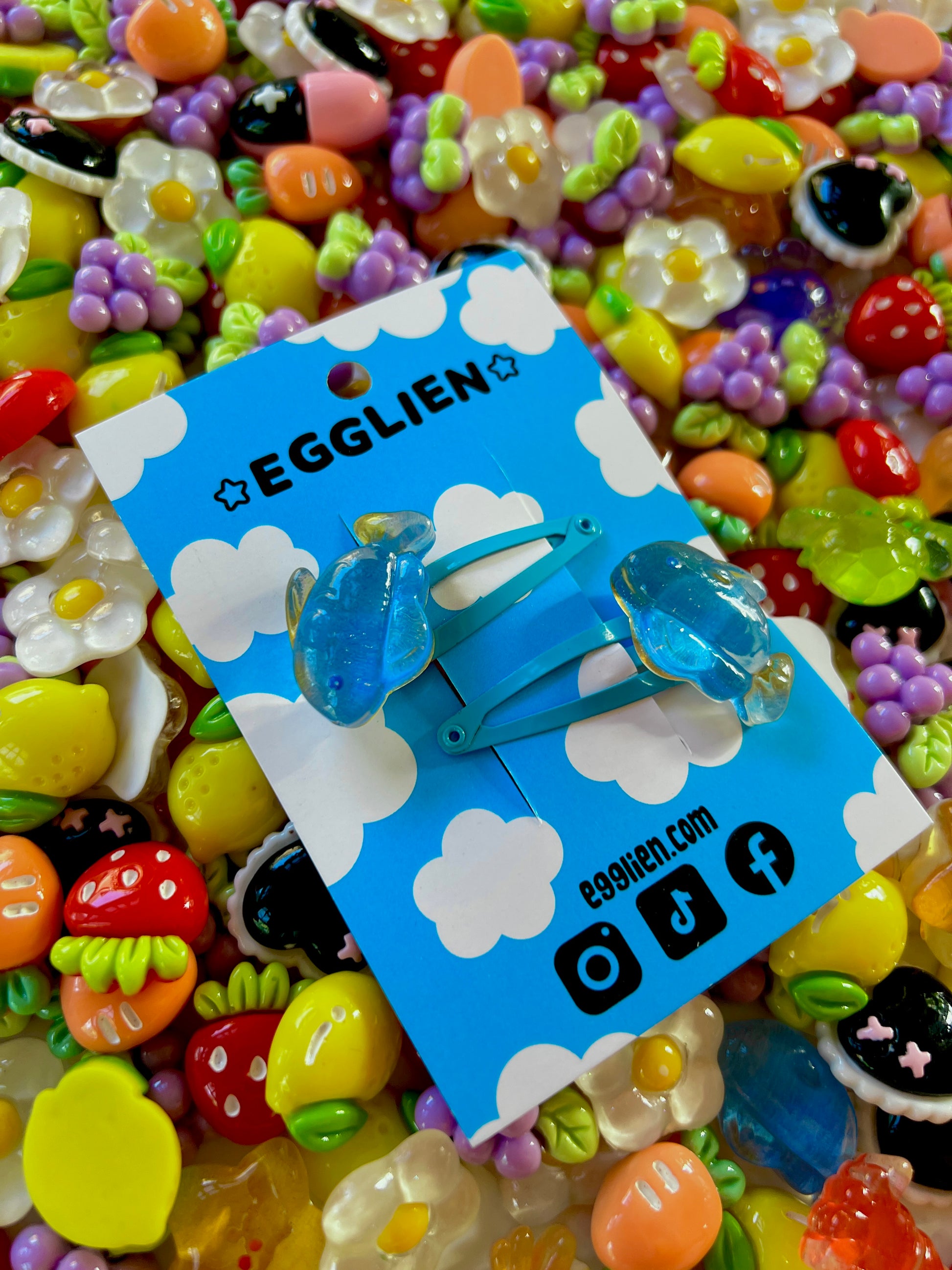 Two blue fish hair clips on a blue card with 'EGGLIEN' branding, surrounded by colorful decorative resin pieces