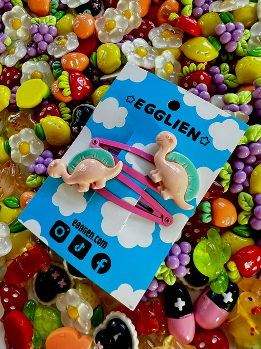 Two peach dinosaurs on bright pink hair-clips on an 'EGGLIEN' card in the center surrounded by colorful decorative items