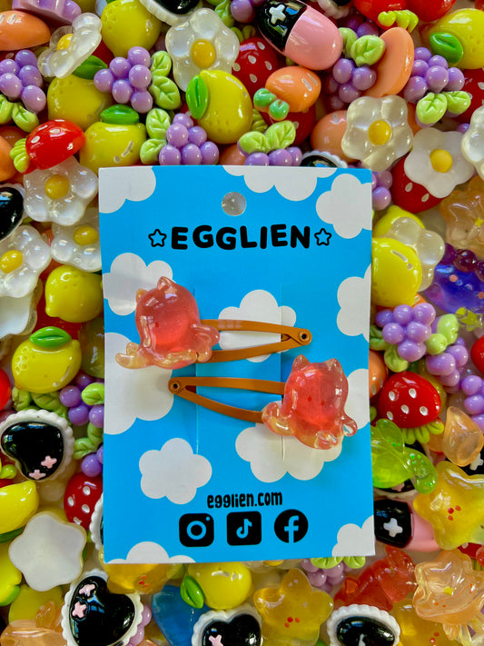 Two red/orange cuttlefish hair clips on a blue card with 'EGGLIEN' branding, surrounded by colorful decorative resin pieces