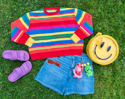 Colorful striped sweater, denim shorts, purple slides, smiley face bag layed flat on grass