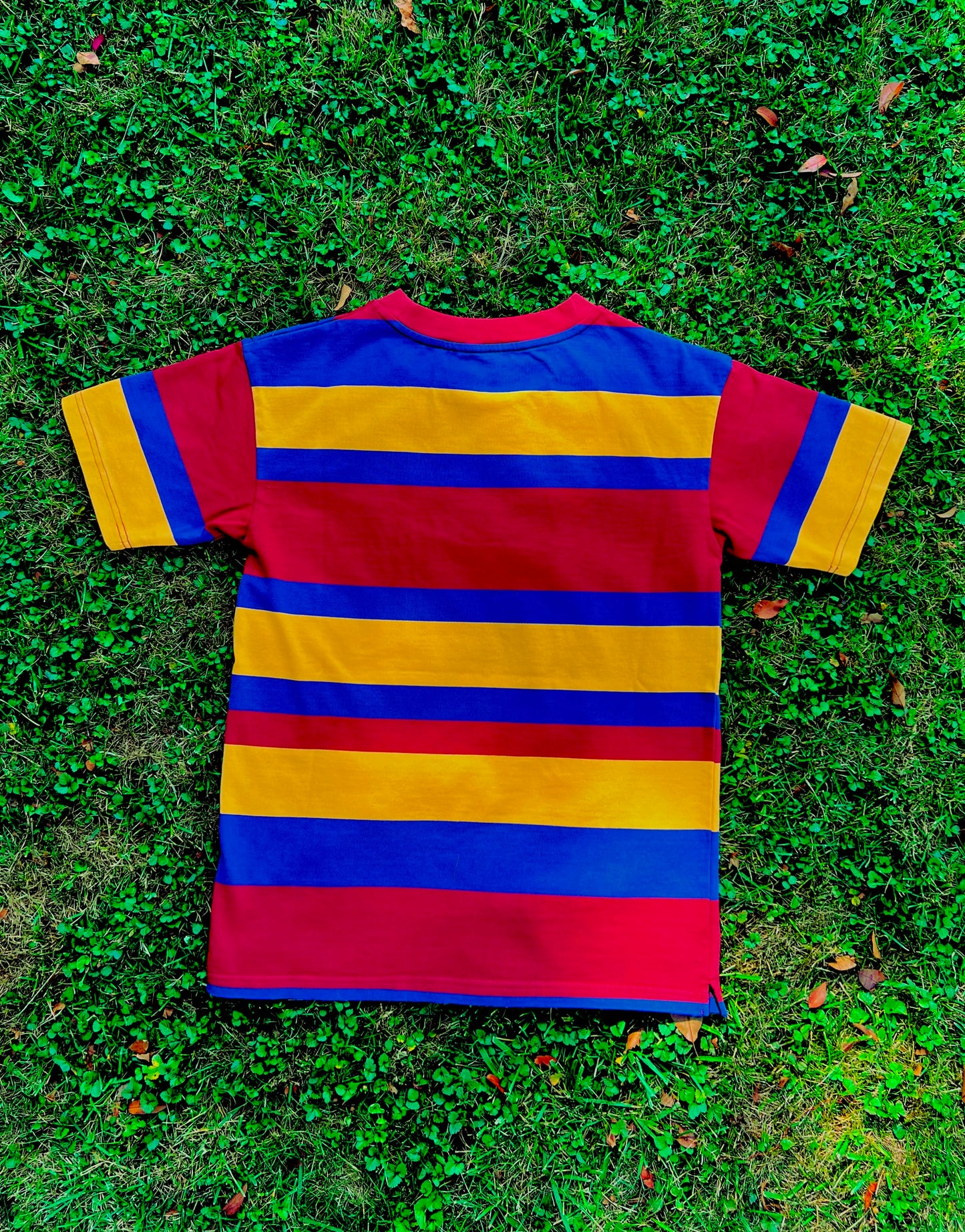 Red, yellow, blue striped shirt with mismatched stripes on the sleeves laying flat and face down on grass