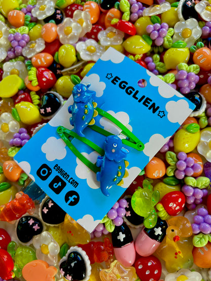 Dark Blue dinosaur-shaped hair clips on a blue card with 'EGGLIEN' branding, surrounded by colorful decorative resin pieces.