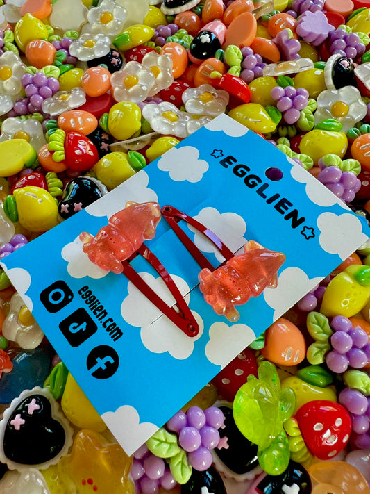 Two red squid hair clips on a blue card with 'EGGLIEN' branding, surrounded by colorful decorative resin pieces