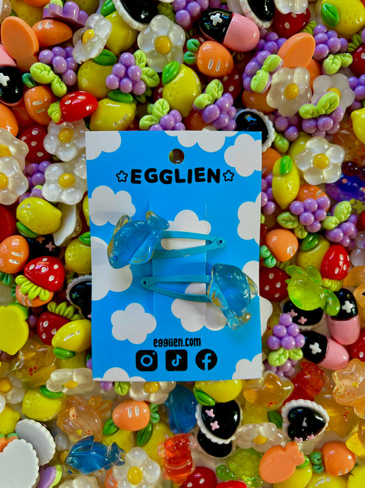 Two blue fish hair clips on a blue card with 'EGGLIEN' branding, surrounded by colorful decorative resin pieces