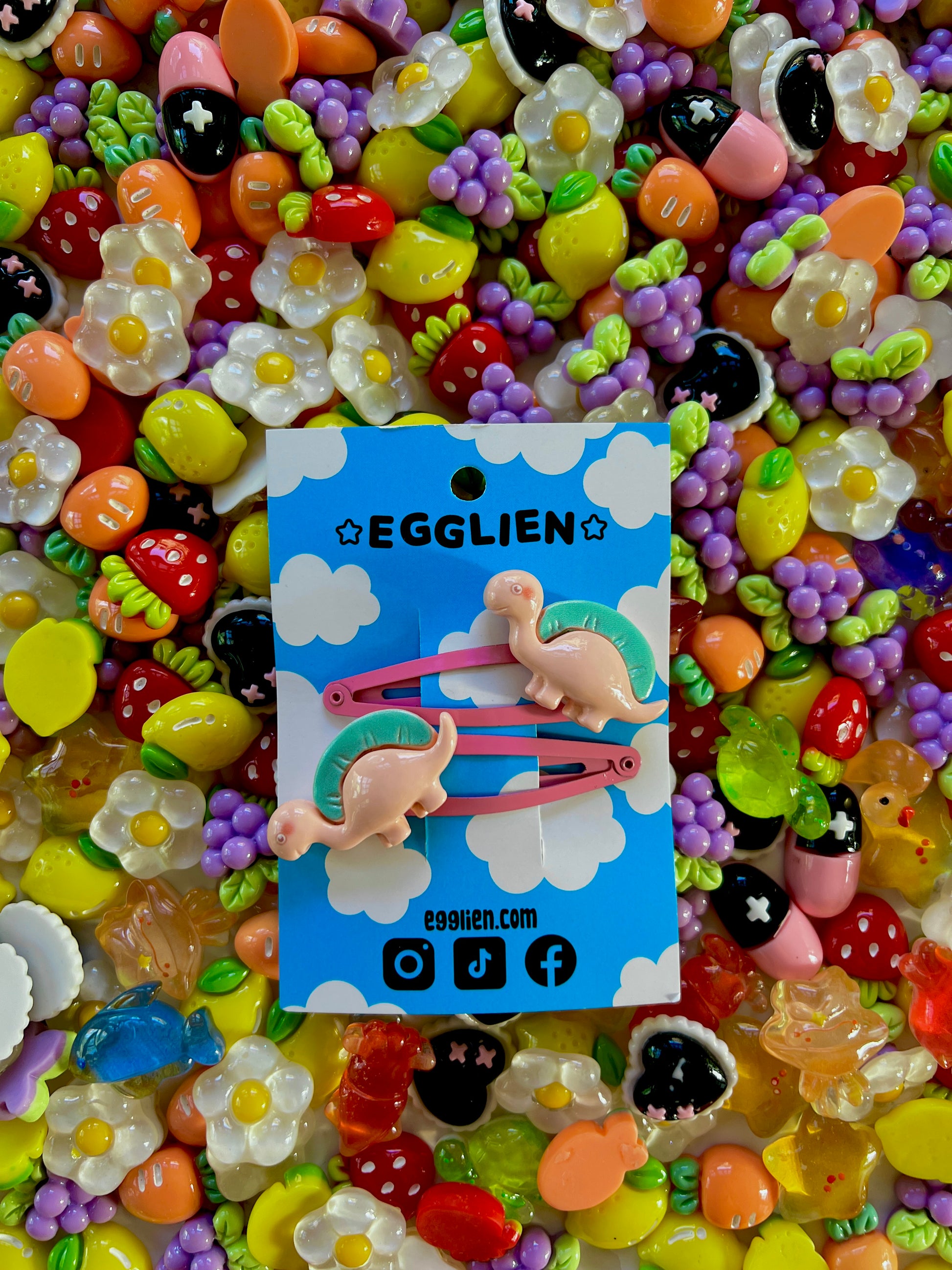Two peach dinosaurs on bright pink hair-clips on an 'EGGLIEN' card in the center surrounded by colorful decorative items